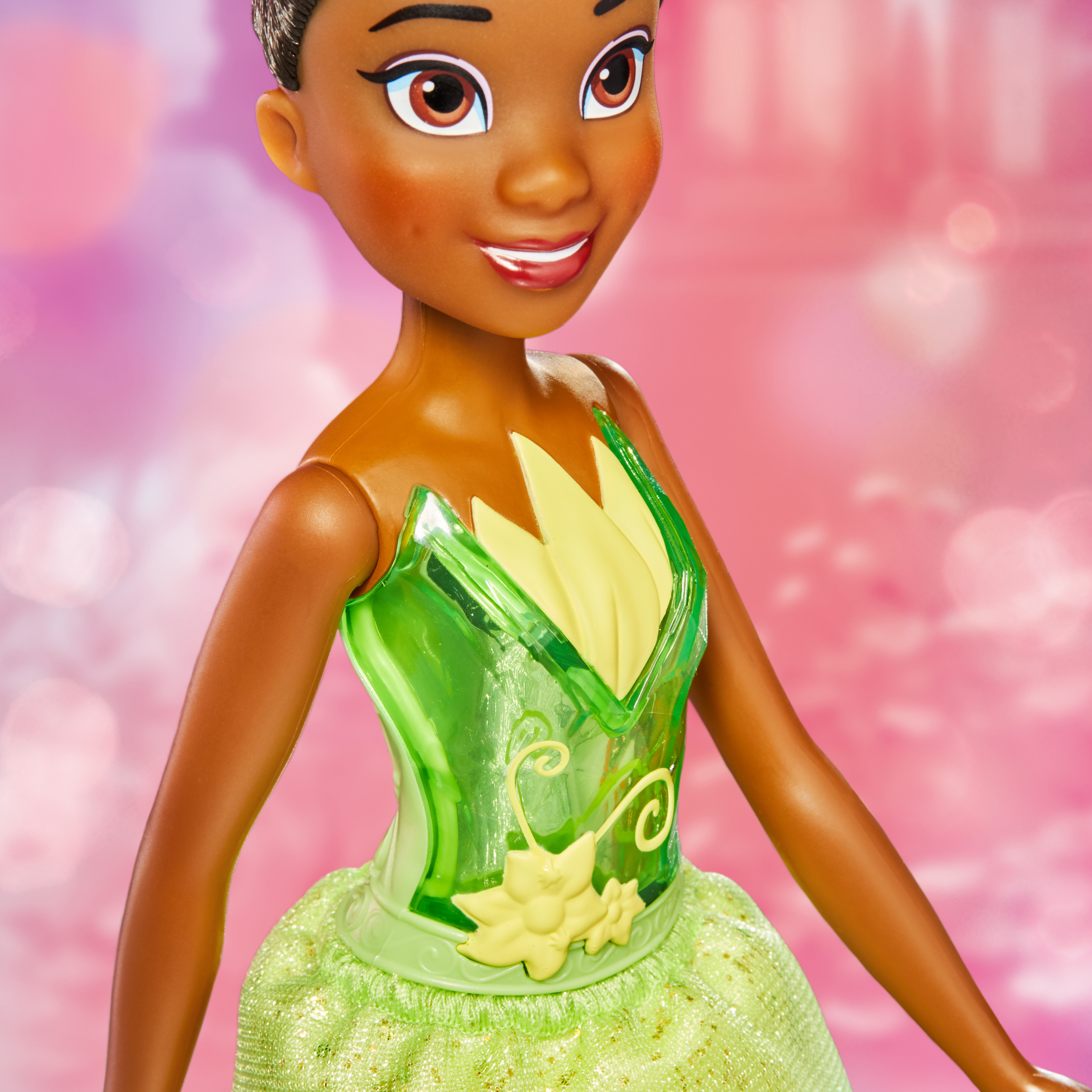 Una bambola di Tiana è centrata, indossa un vestito verde. Lo sfondo è rosa. La sua pelle è marrone e sorride.