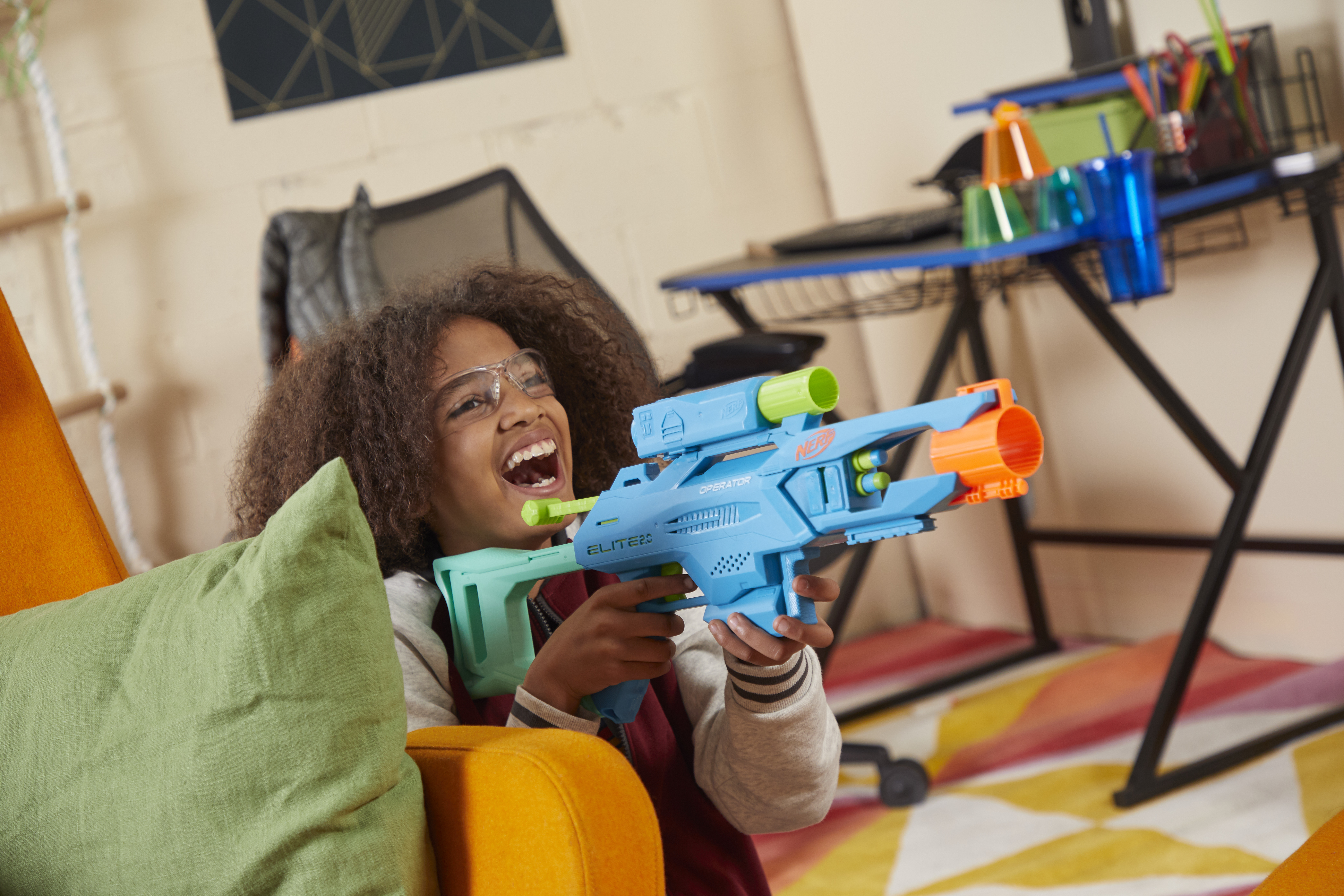 Dziecko w okularach uśmiecha się, trzymając niebieski pistolet Nerf. Widoczne są zielone i pomarańczowe akcenty. W tle biurko i dywan.