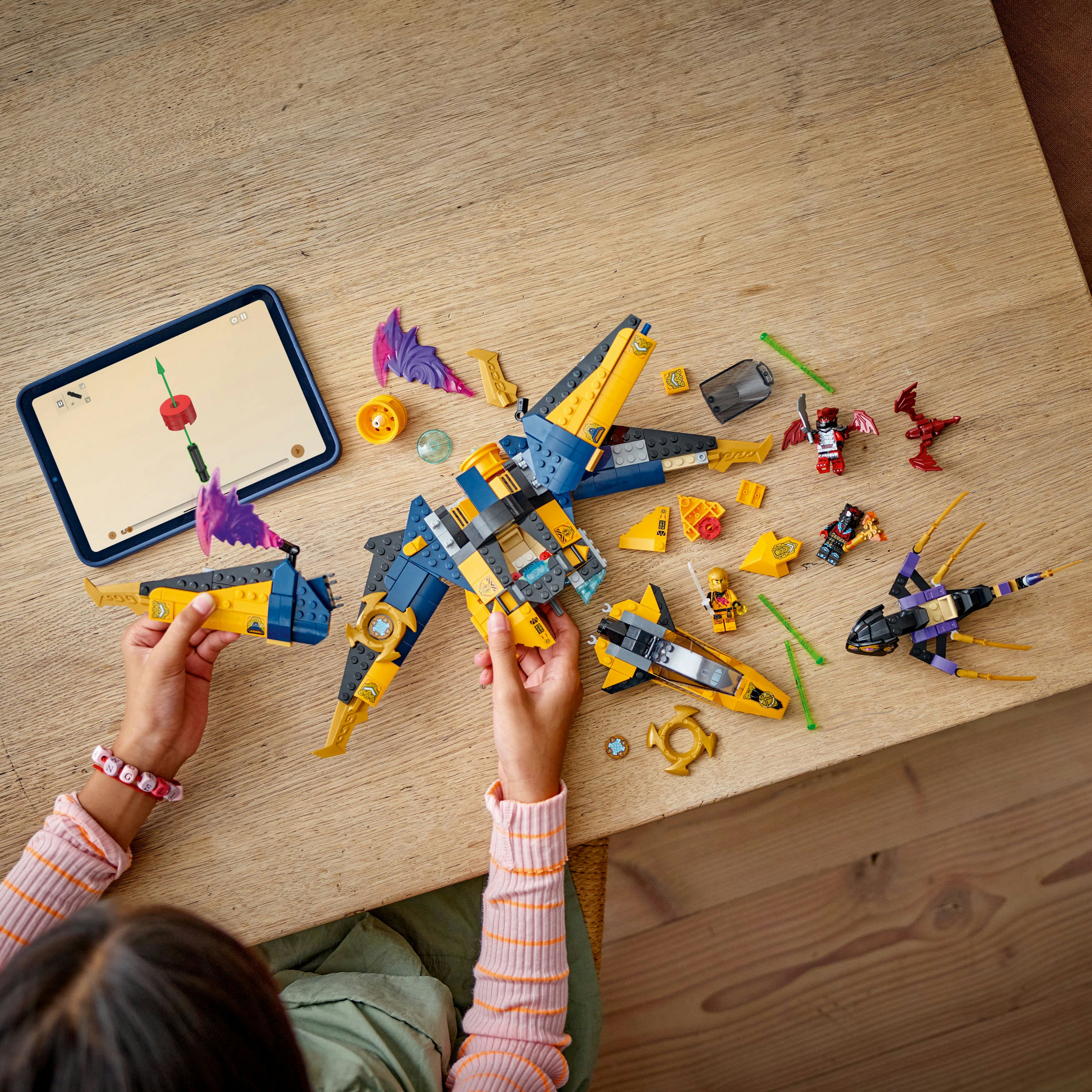 Dziecko składające zestaw Lego na drewnianym stole. Zestaw zawiera odrzutowiec, minifigurki i tablet z instrukcjami.