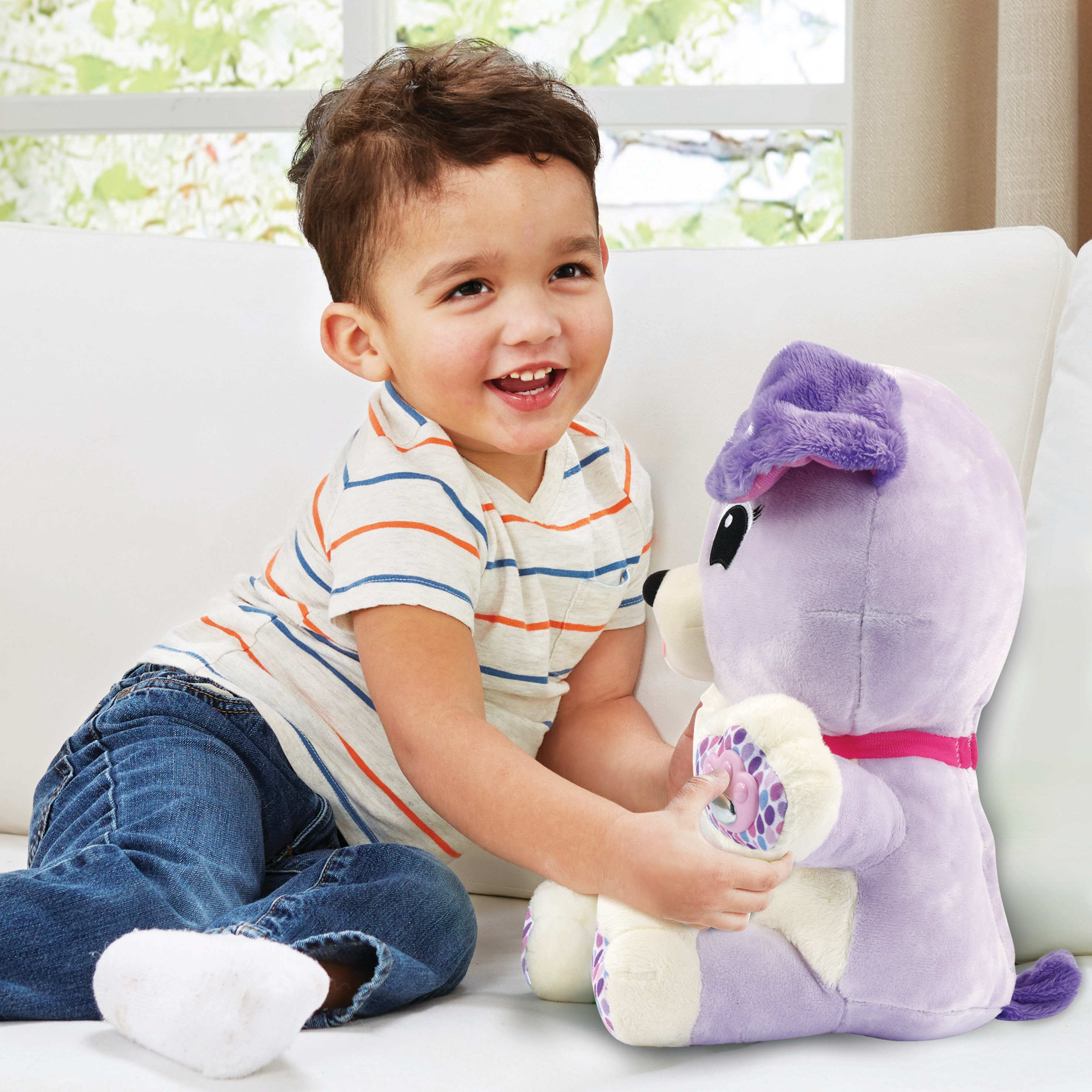 Un garçon souriant en jean tient une peluche de chien violet sur un canapé blanc. Une fenêtre avec un feuillage vert est en arrière-plan.