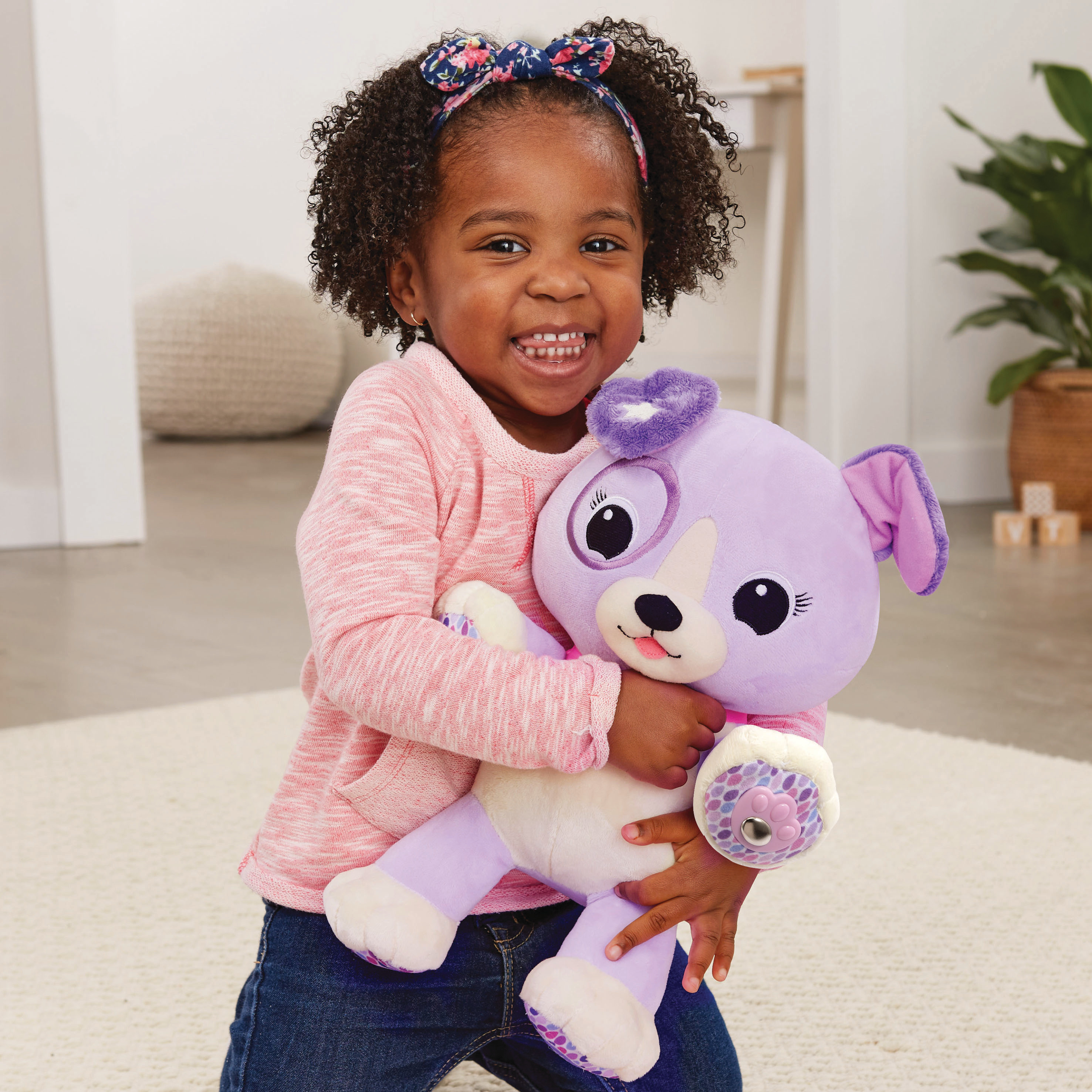 Une fillette noire souriante tient un chiot en peluche violet. Elle porte un haut rose, un jean et un serre-tête floral.