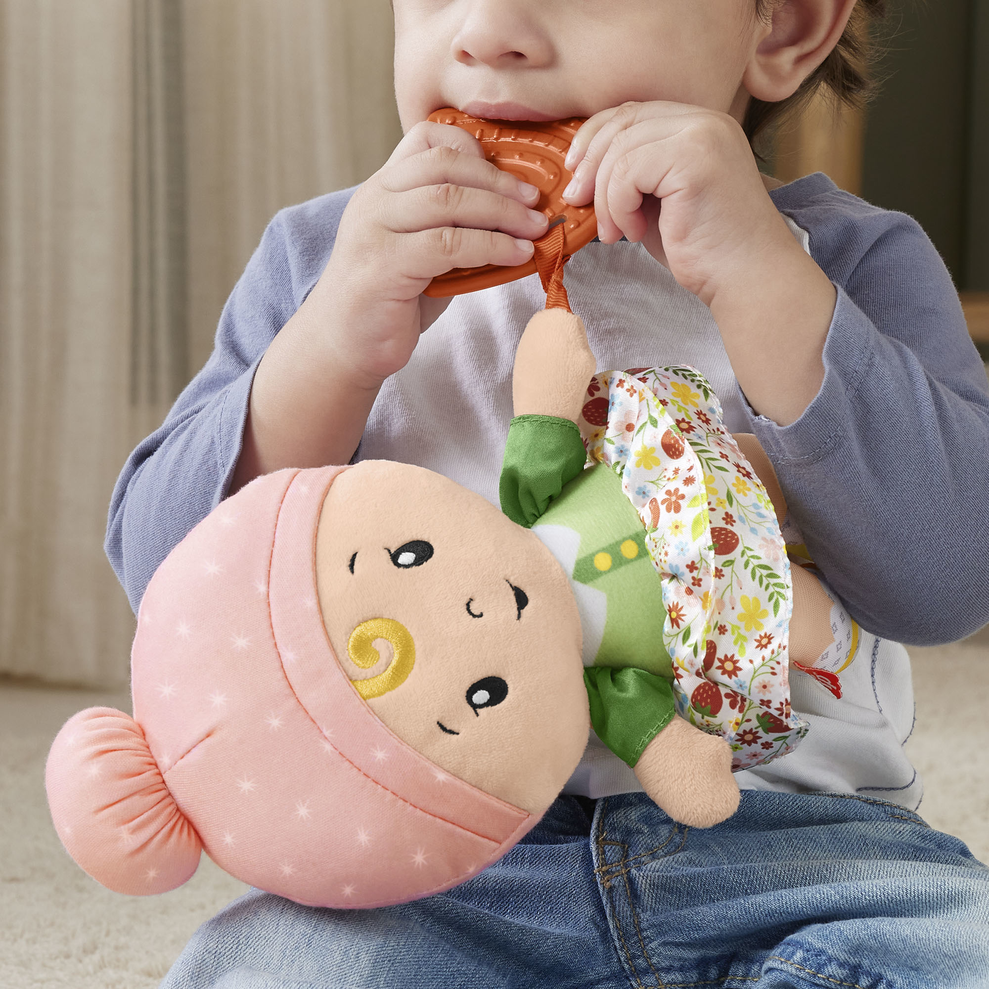 Dziecko trzyma i gryzie pomarańczową zabawkę do ząbkowania. Lalka z różową głową jest przed dzieckiem.