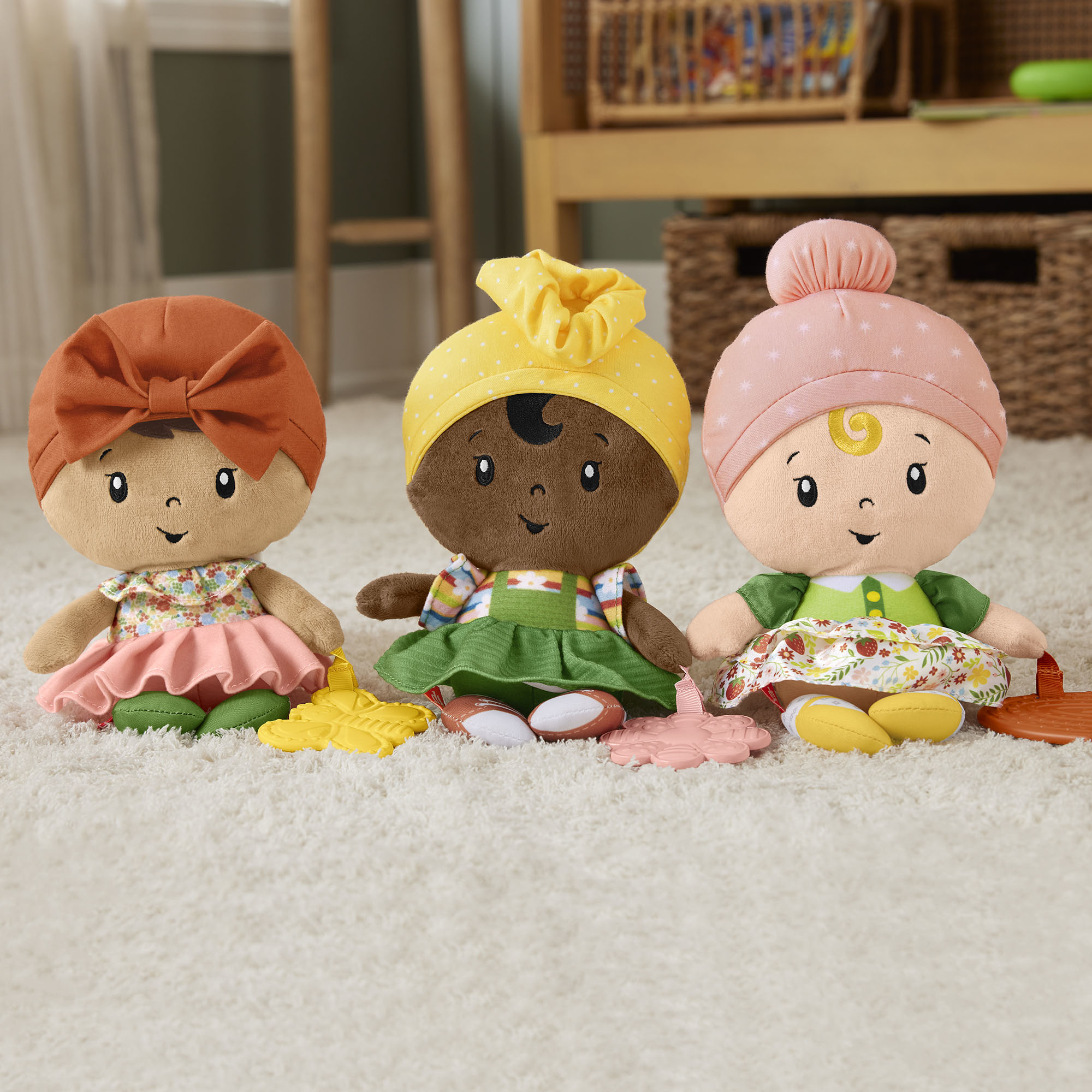 Trzy pluszowe lalki o różnych odcieniach skóry i nakryciach głowy siedzą na białym dywanie z zabawkami.