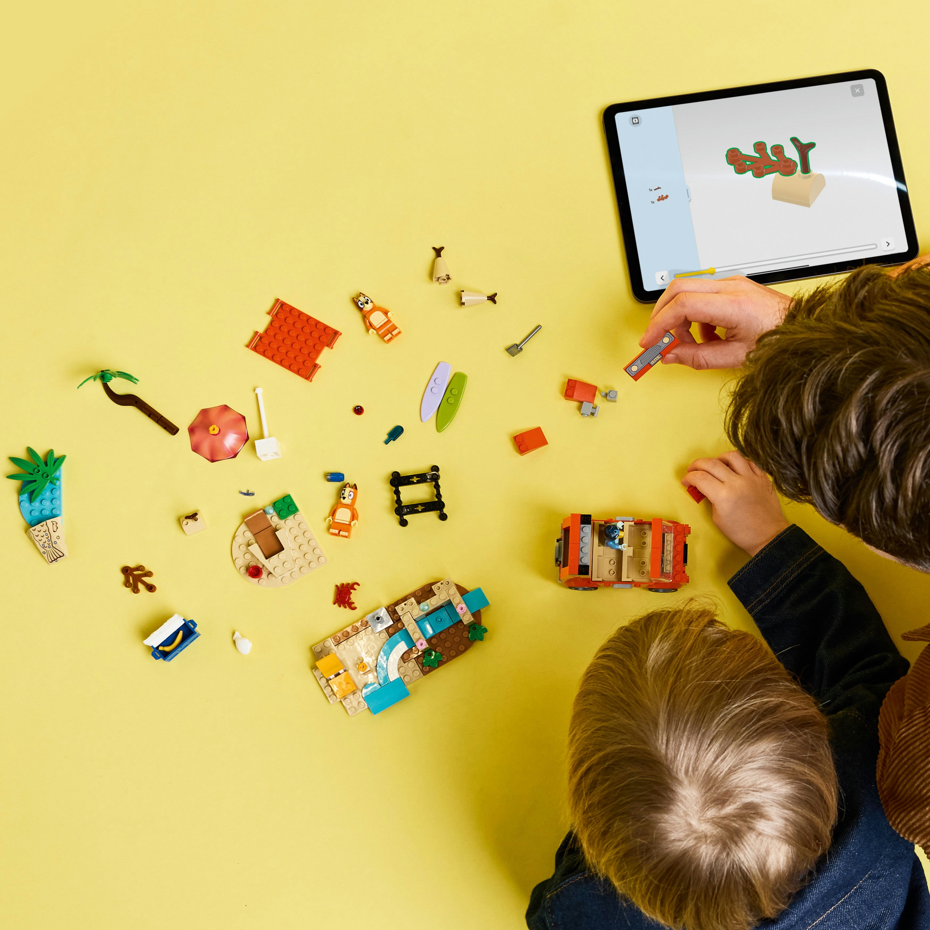 Des enfants construisent avec des pièces Lego sur une surface jaune, avec des instructions sur une tablette. La scène est vue d'en haut.