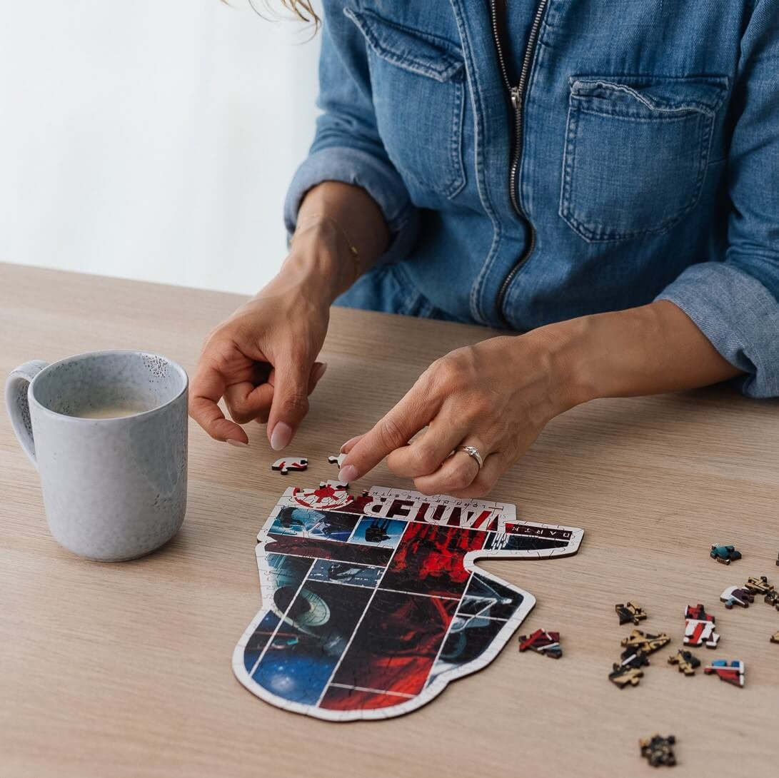 Una persona sta assemblando un puzzle di Darth Vader su un tavolo di legno. Una tazza di caffè è a sinistra. La persona indossa una camicia di jeans.