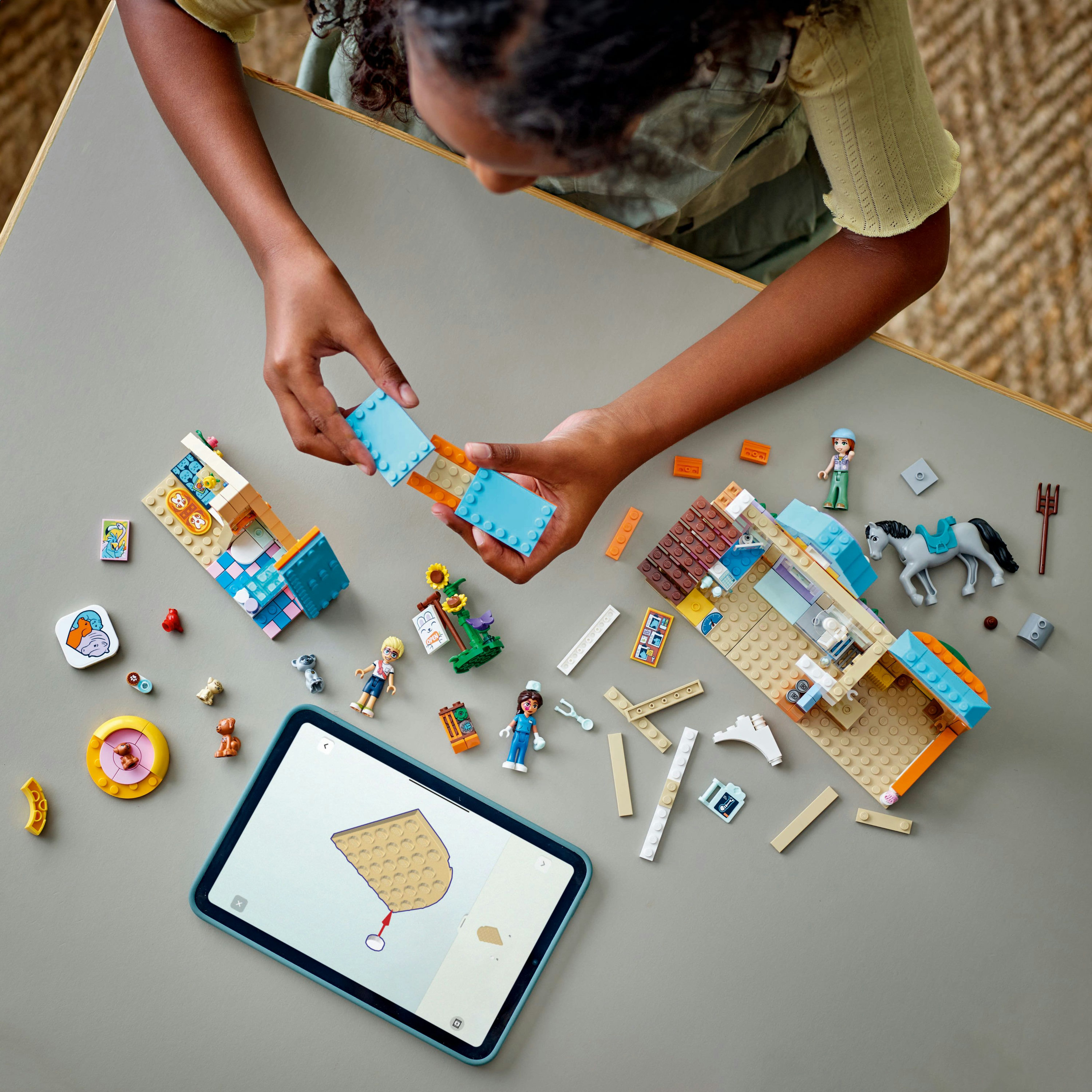Osoba składająca klocki LEGO. Niebieskie i pomarańczowe konstrukcje otaczają tablet z instrukcjami. Obecne są minifigurki i koń.