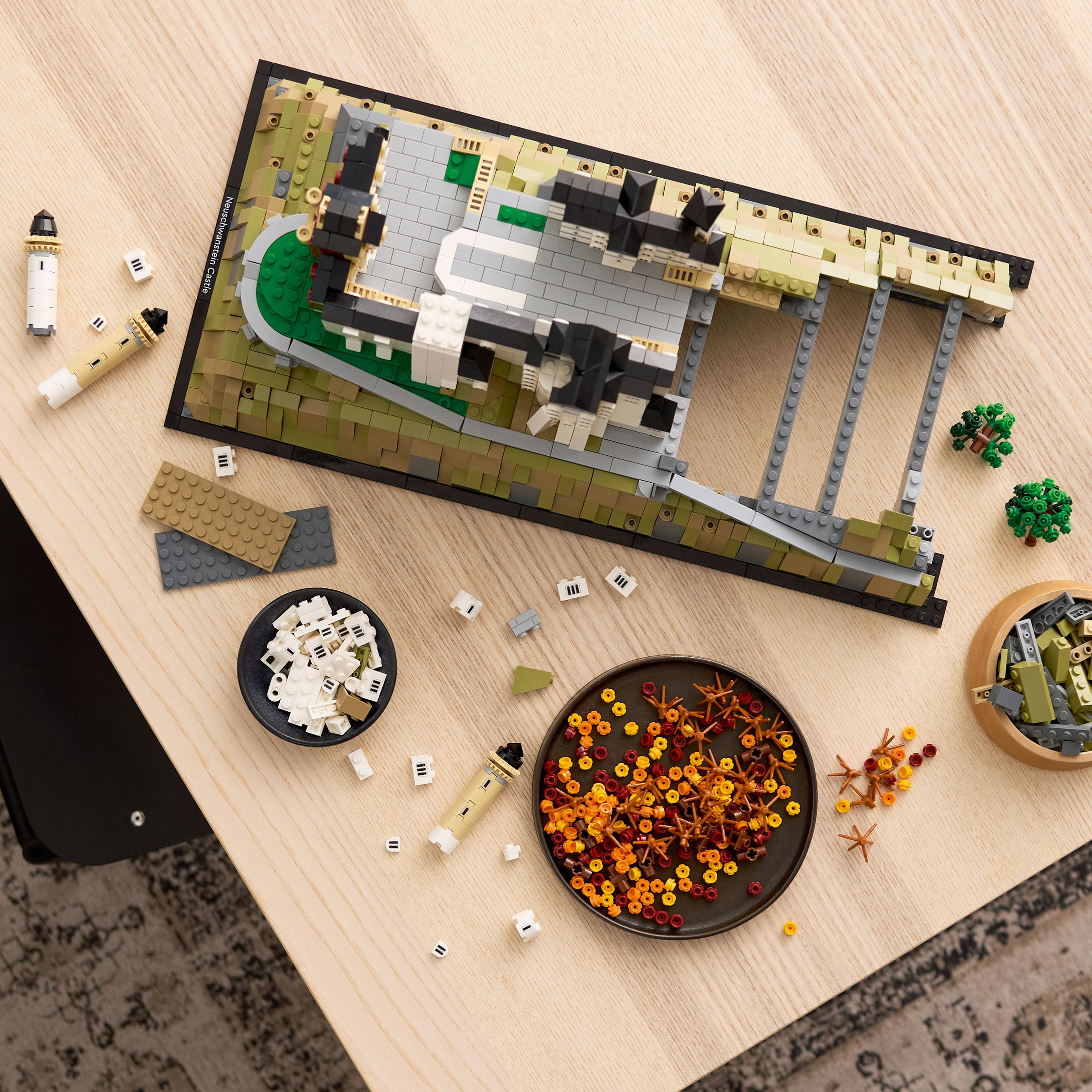 Model LEGO zamku na stole z rozrzuconymi częściami i małymi drzewami. Widoczne są również latarnie morskie i małe miski.