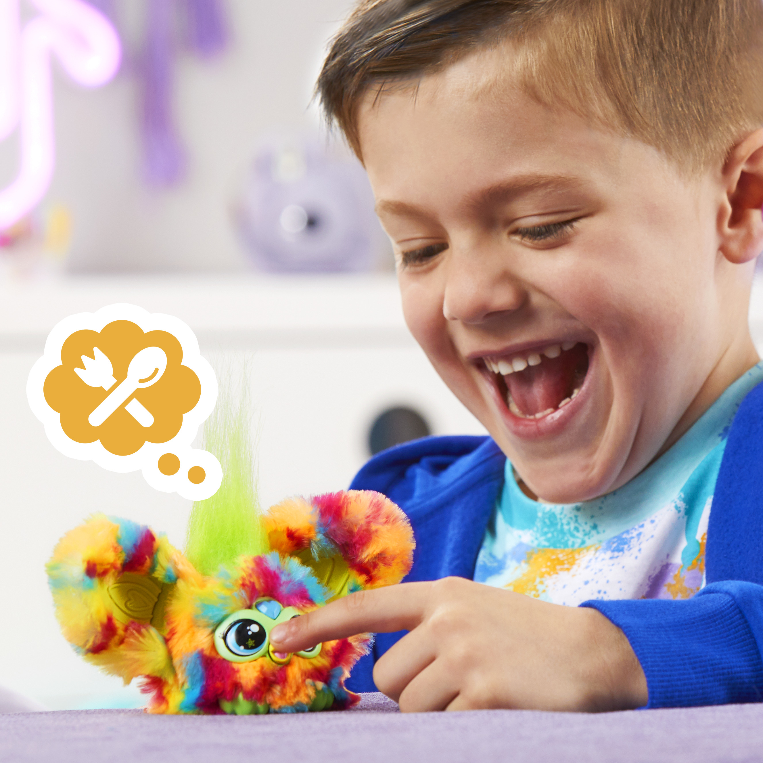Un niño se ríe mientras toca un juguete colorido. Una burbuja de pensamiento muestra un tenedor y una cuchara.