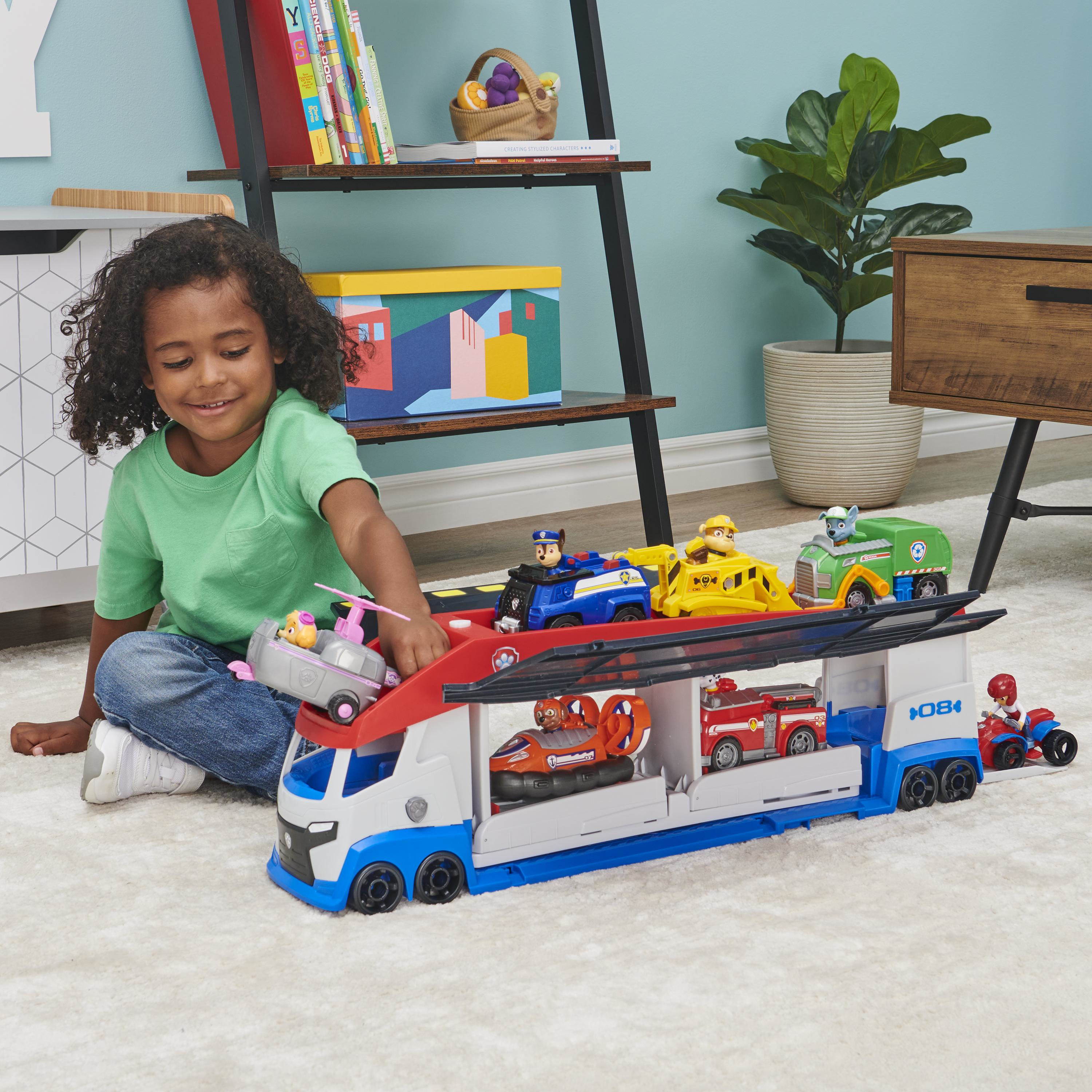 Ein Kind spielt auf einem Teppich mit einem Paw Patrol-Spielzeug-LKW, der mit verschiedenen Charakterfahrzeugen gefüllt ist.