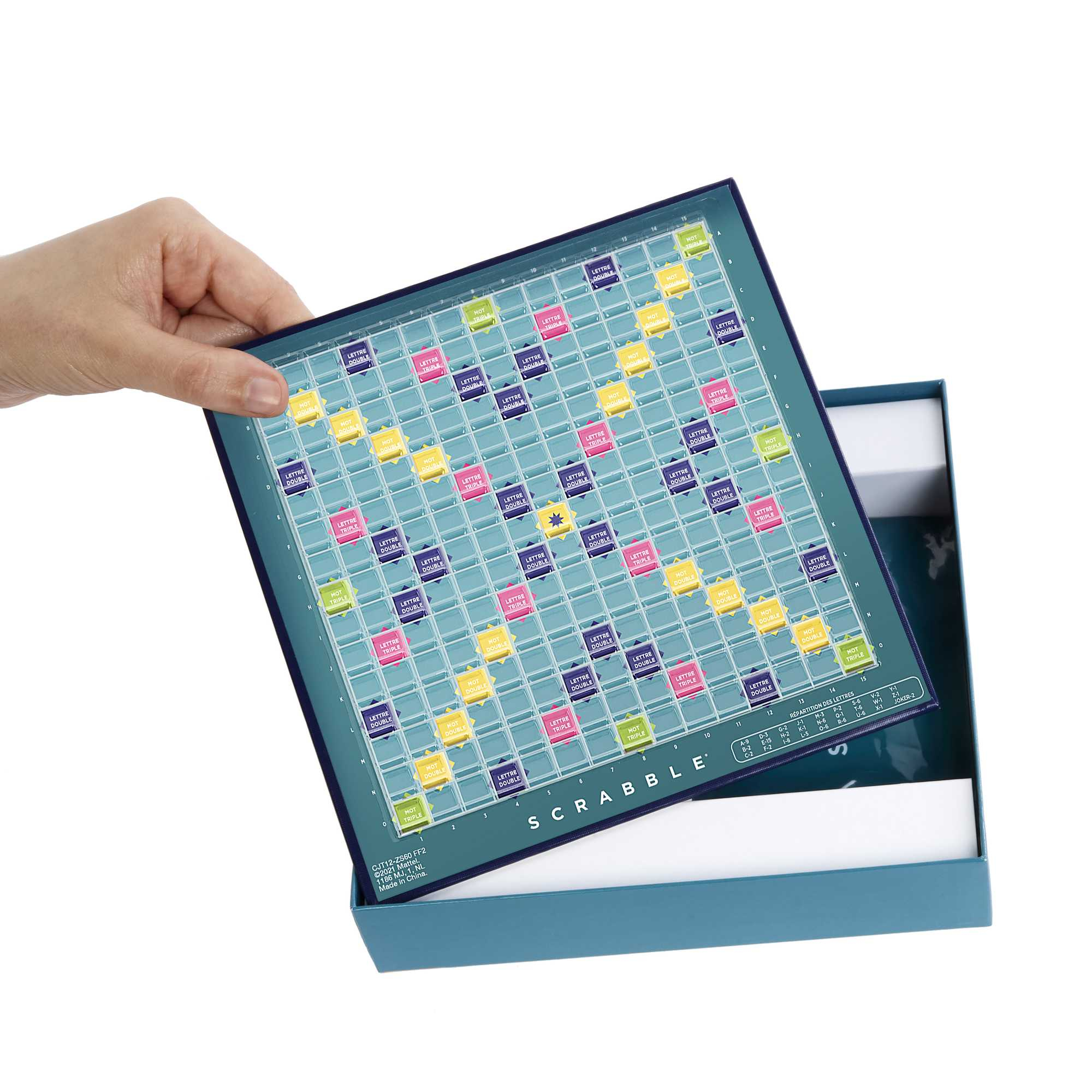 Een hand houdt een Scrabble-bord boven een open doos. Het bord is blauw en groen met letter- en woordvakken.