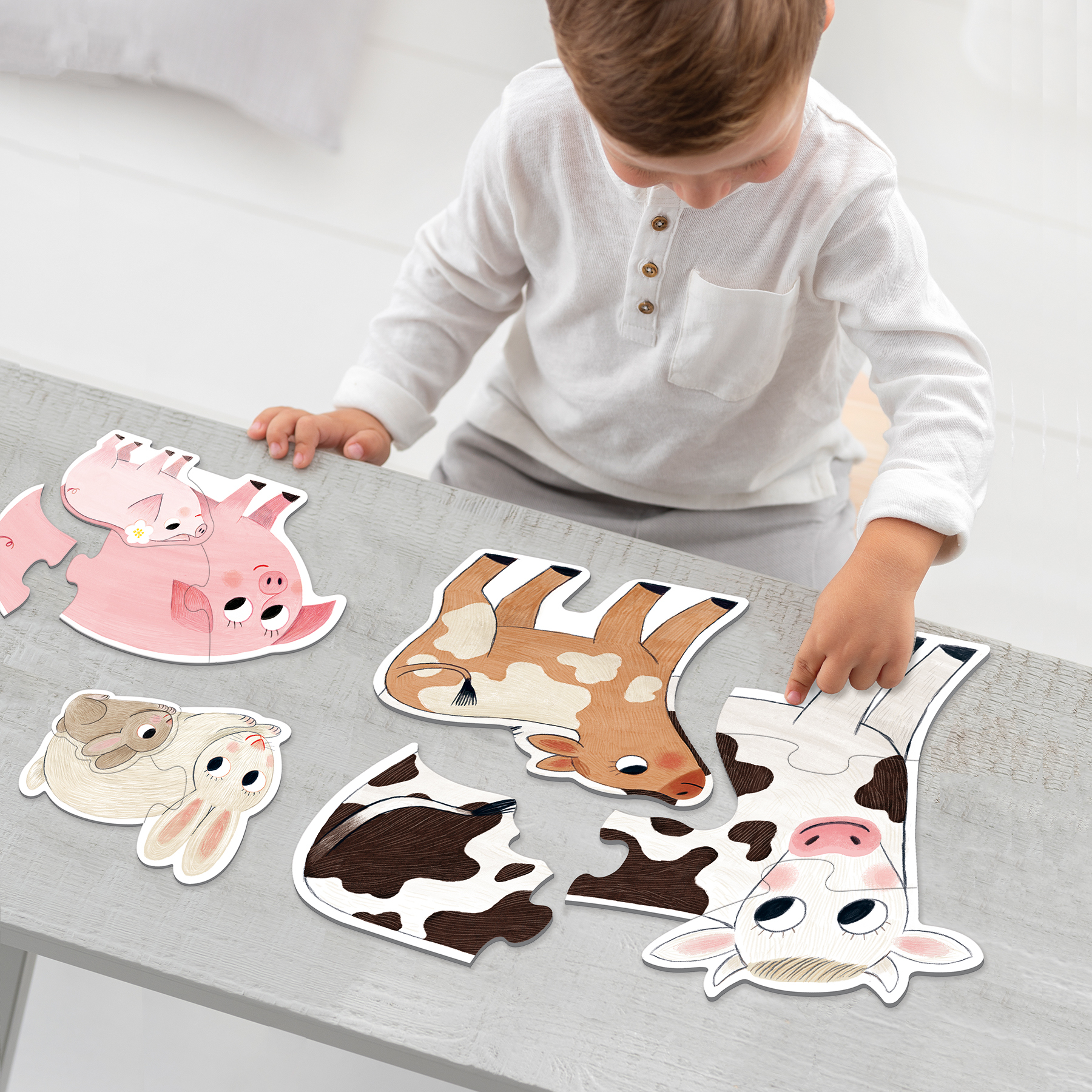 Dziecko bawi się puzzlami ze zwierzętami na szarym stole. Widoczne są puzzle z świniami, krowami i królikami.