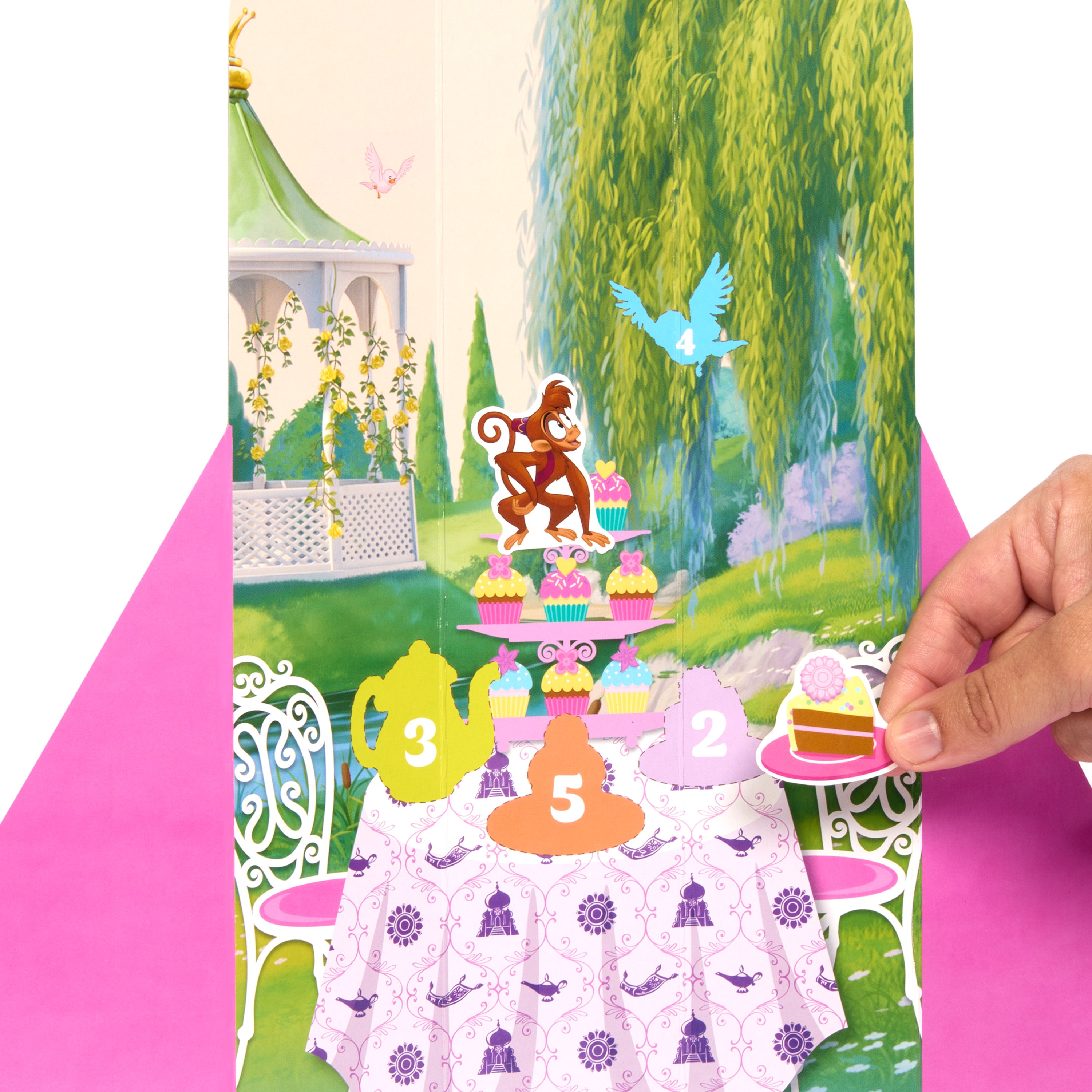 Karta pop-up z małpą, babeczkami i elementami przyjęcia herbacianego. Ręka trzymająca ciasto. Tło ma scenę ogrodu.