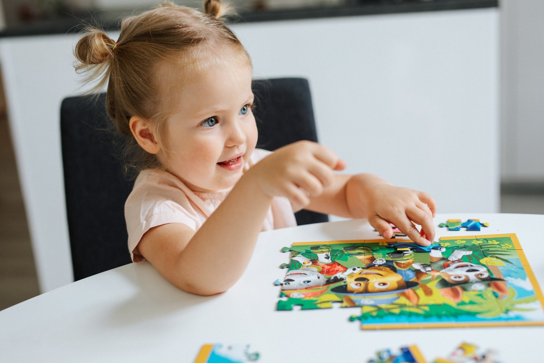 A smiling toddler with pigtails is doing a puzzle at a table. The puzzle has cartoon characters.