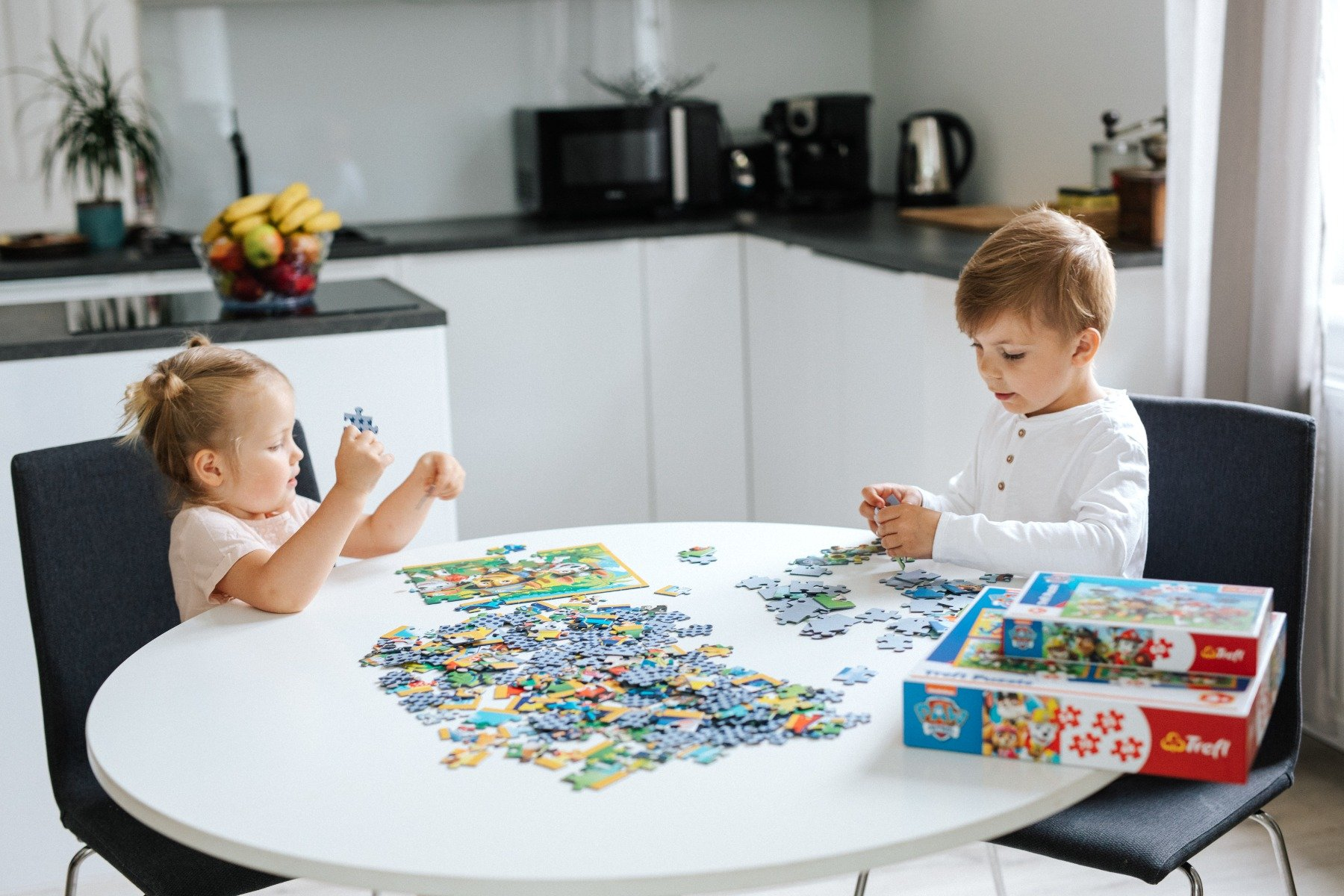 Two children assemble a puzzle at a white table. A bowl of fruit and kitchen appliances are in the background.