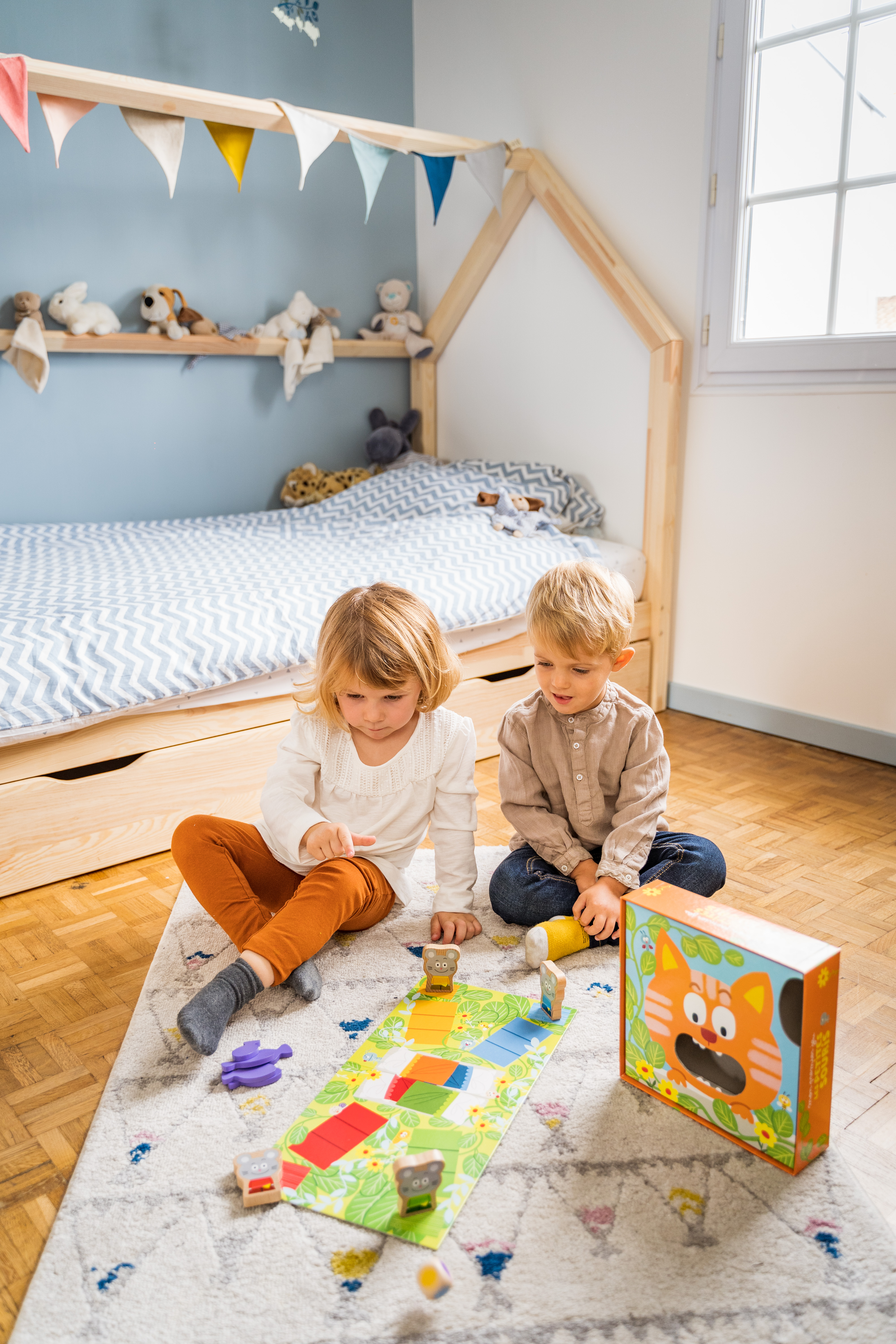 Twee kinderen spelen een bordspel op een kleed. Een bed en een raam op de achtergrond. Speelgoed op een plank.