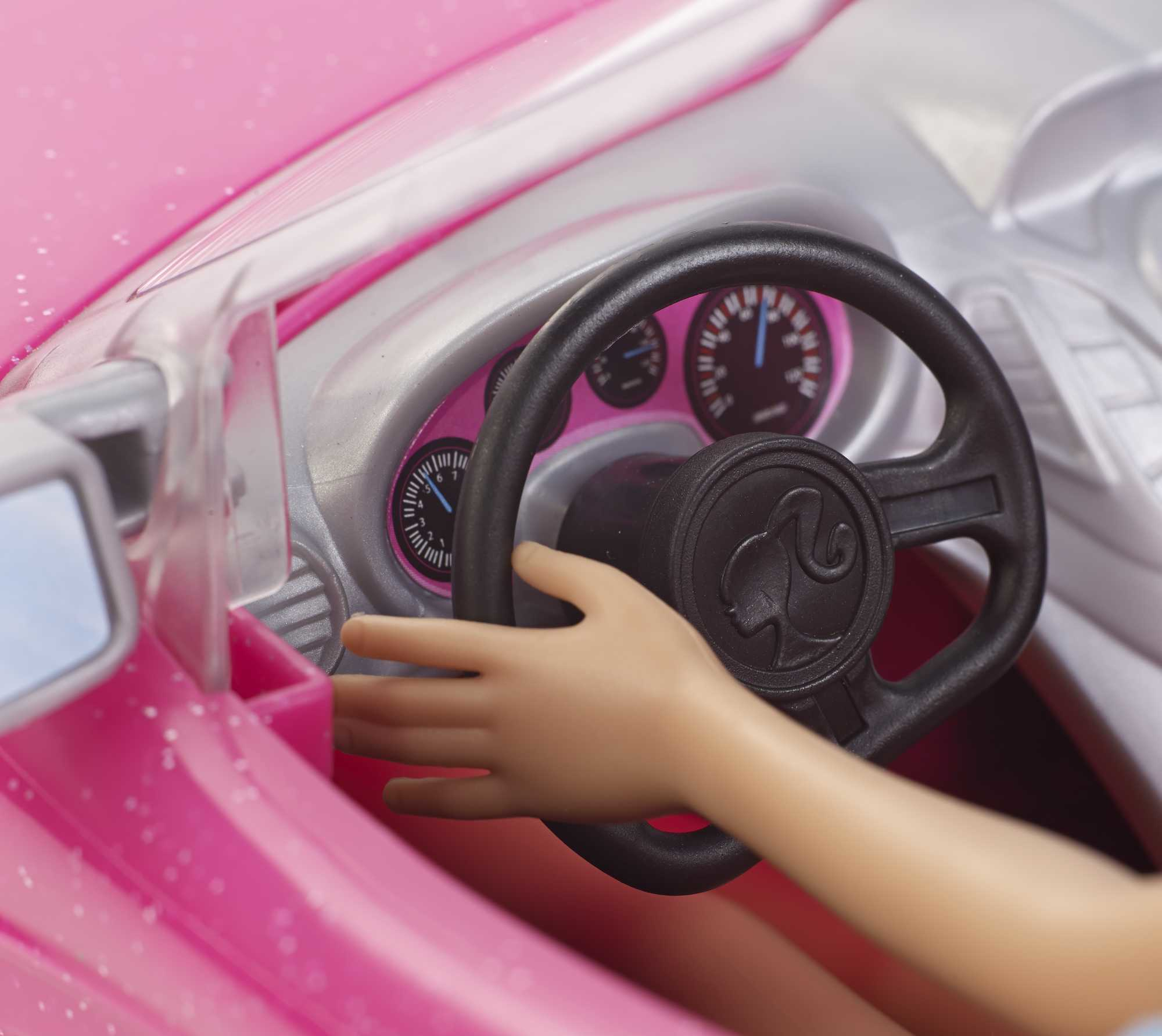 Zbliżenie dłoni lalki na czarnej kierownicy z logo Barbie, wewnątrz różowej deski rozdzielczej.