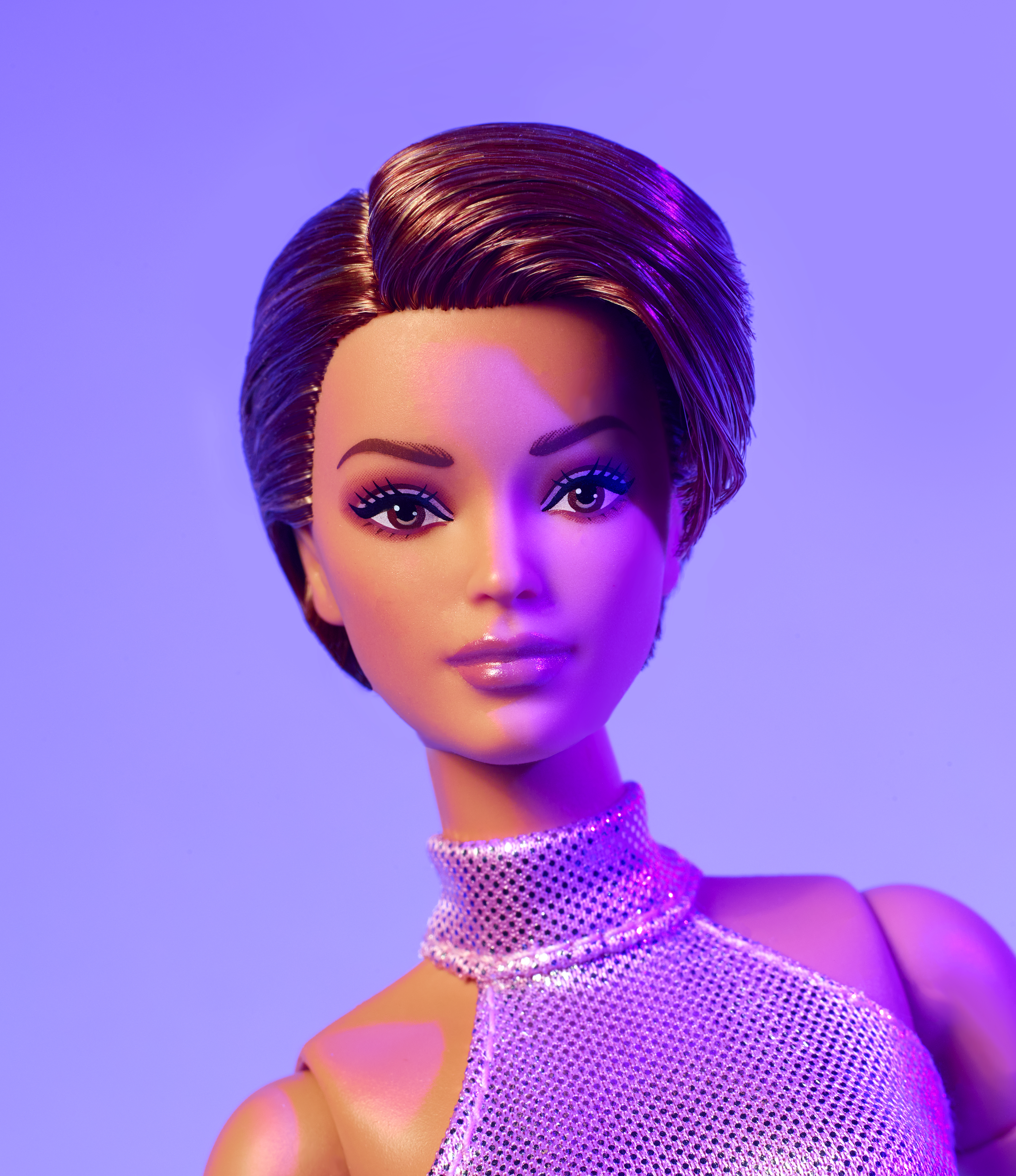 Eine Puppe mit braunem Haar und Make-up, die ein glitzerndes Oberteil trägt, vor einem lilafarbenen Hintergrund.