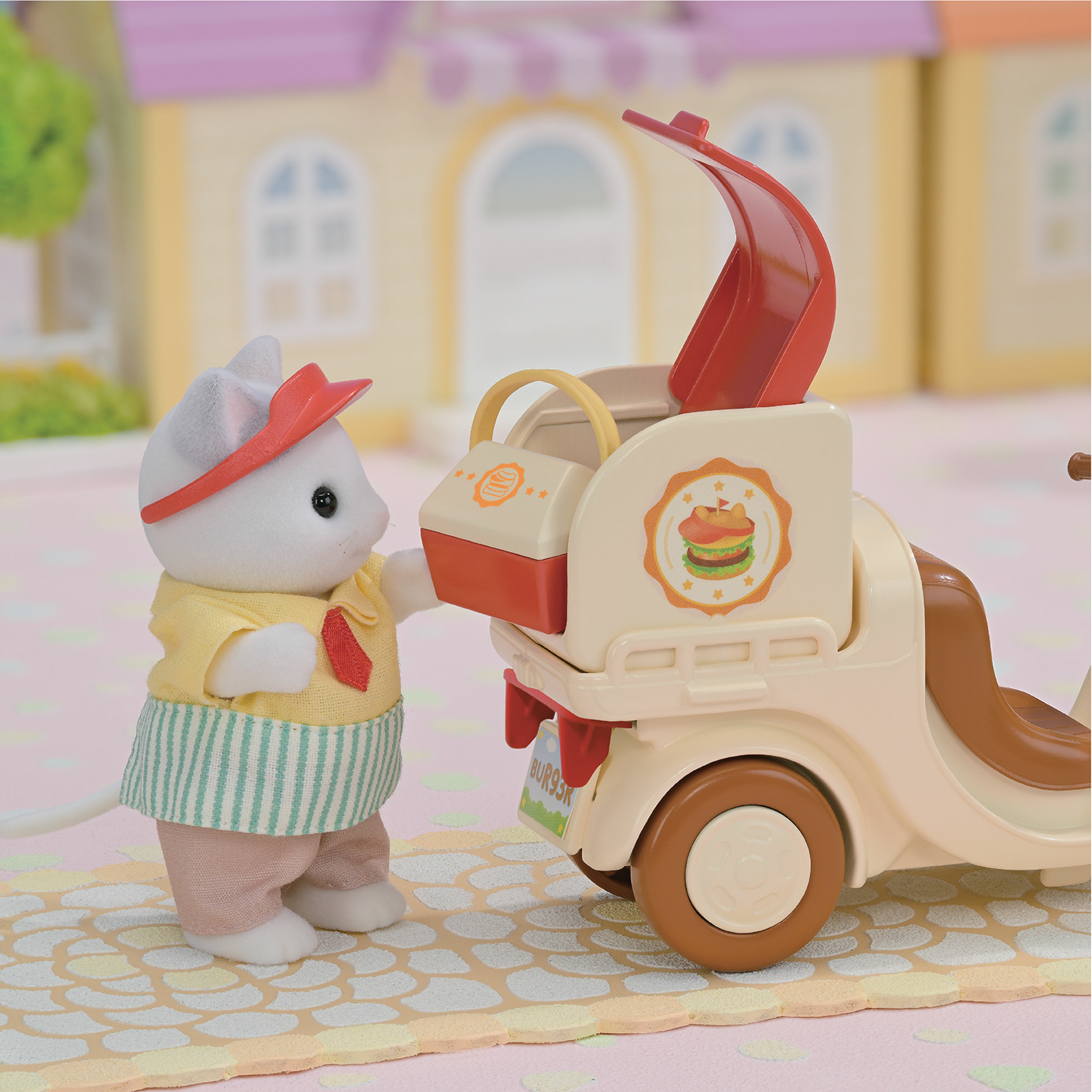 Una figura de ratón blanco con camisa y delantal amarillos junto a un carrito de hamburguesas beige. Un edificio al fondo.