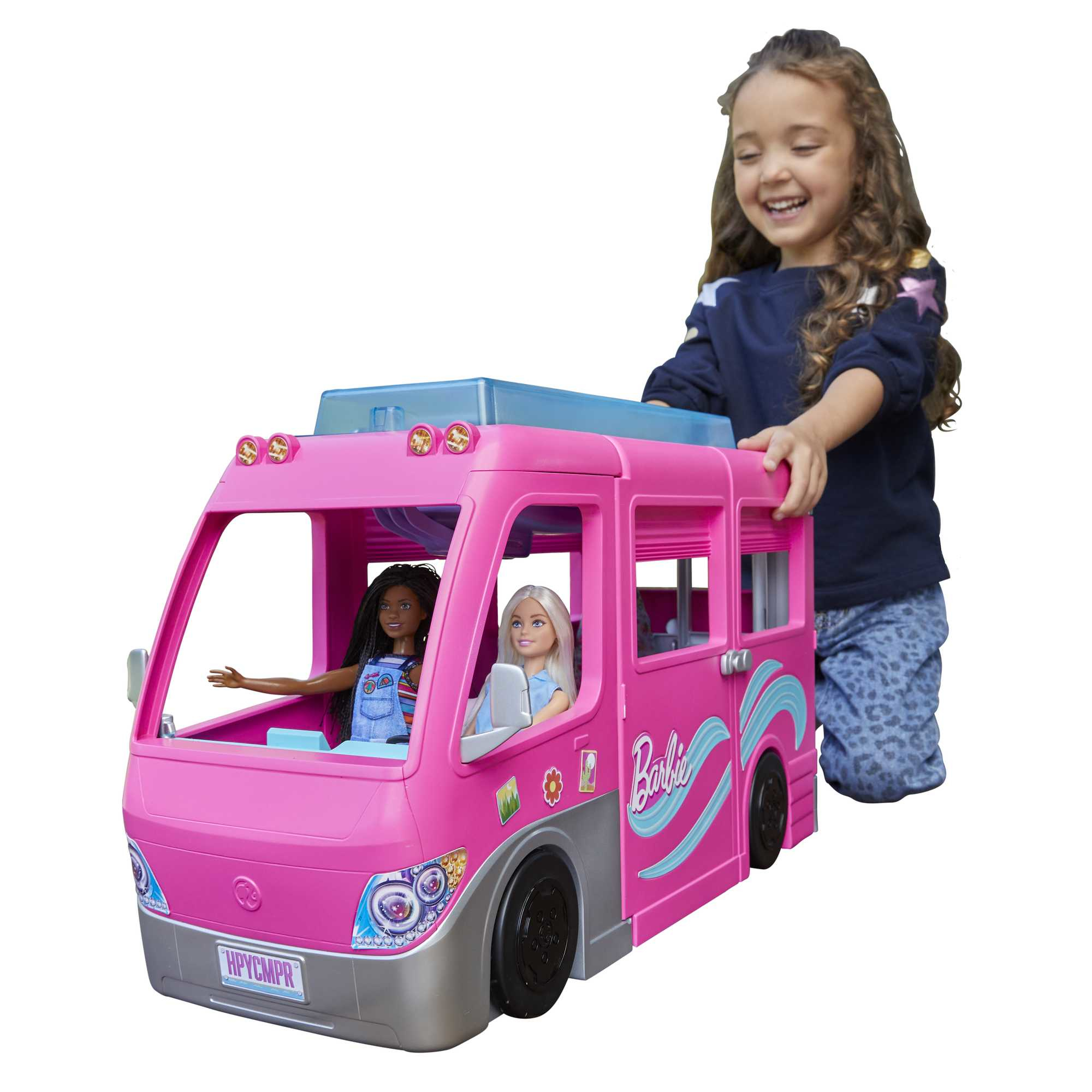 Una bambina gioca con un camper Barbie rosa, con bambole dentro. La bambina sorride, toccando il camper.