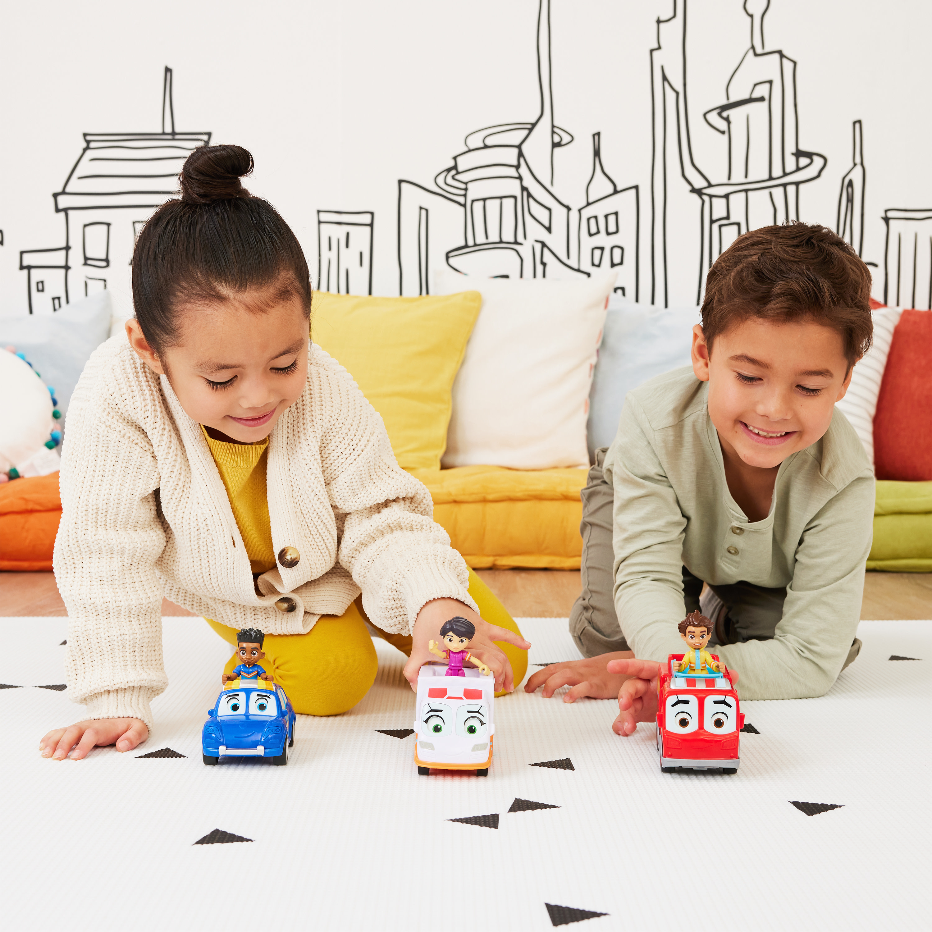 Dwoje dzieci bawi się samochodzikami. Dziewczynka i chłopiec siedzą na białym dywanie z czarnymi trójkątami. Na ścianie rysunek miasta.