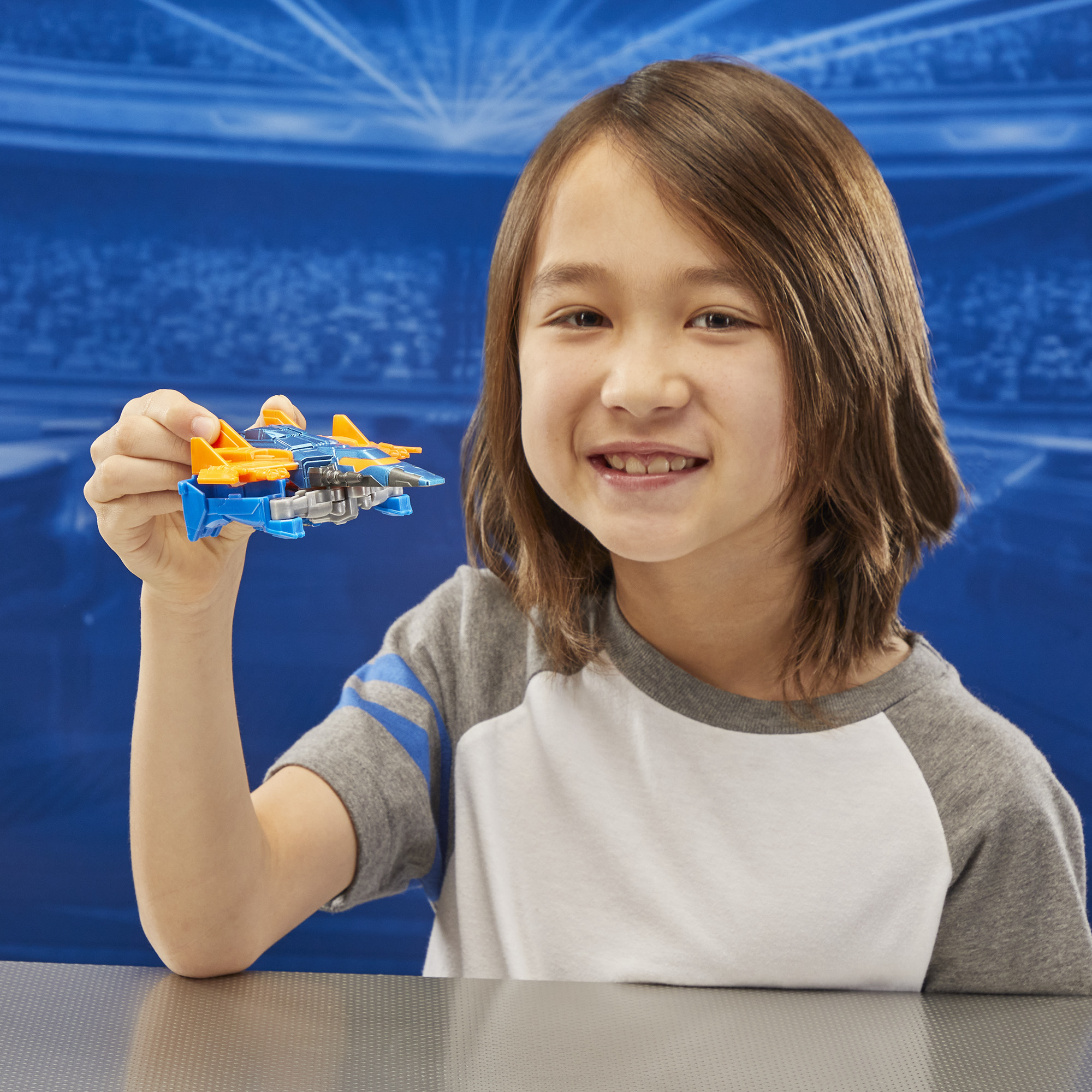 Chłopiec trzyma niebiesko-pomarańczowy statek kosmiczny, uśmiechając się. Tło to rozmyty niebieski stadion.