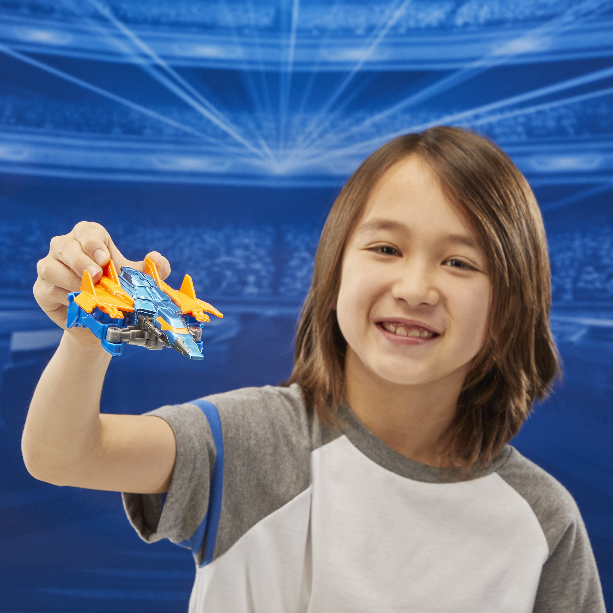 Uśmiechnięte dziecko trzyma niebiesko-pomarańczowy pojazd-zabawkę. Tło to niebieska grafika stadionu.