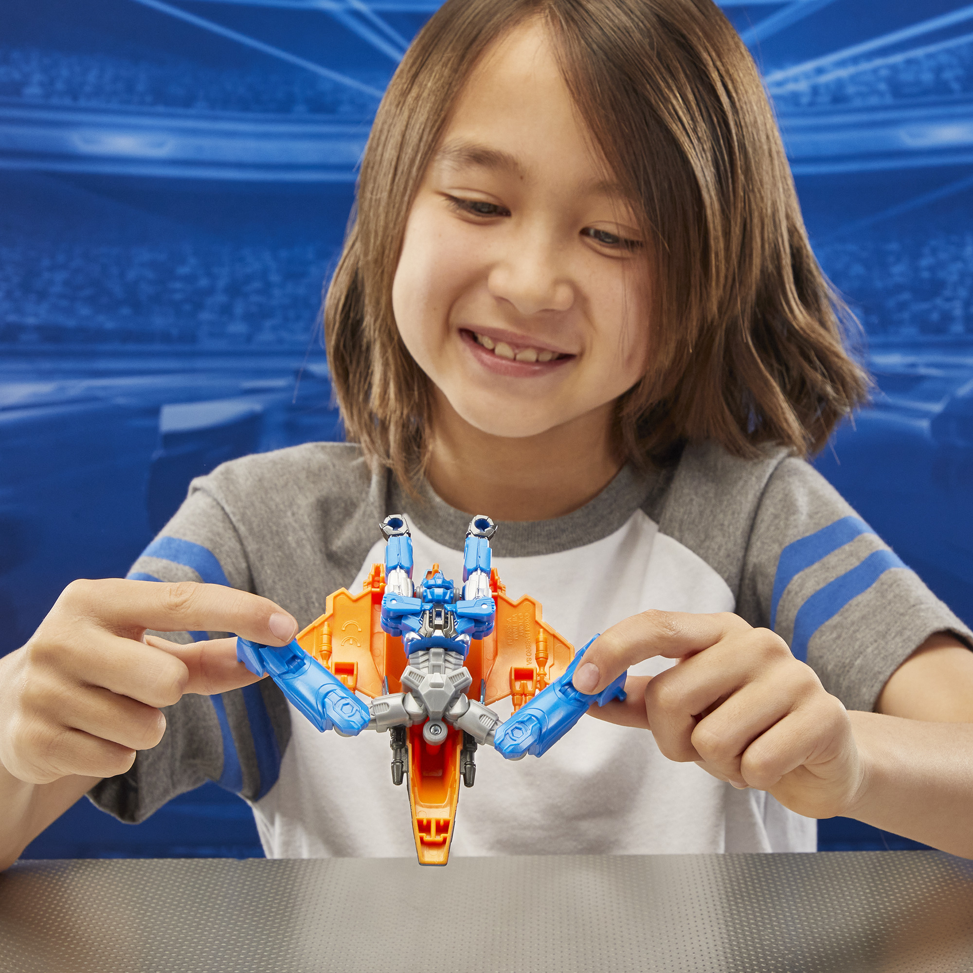 Chłopiec trzyma zabawkę samolotu. Samolot jest pomarańczowy, niebieski i srebrny. Uśmiecha się. Niebieskie tło.