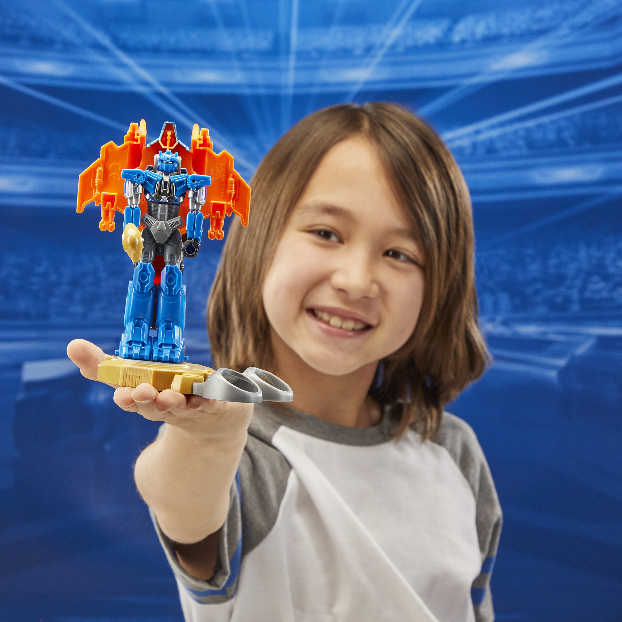 Dziecko trzyma niebiesko-pomarańczowego robota. Dziecko się uśmiecha. Za nim niebieskie tło z białymi liniami.