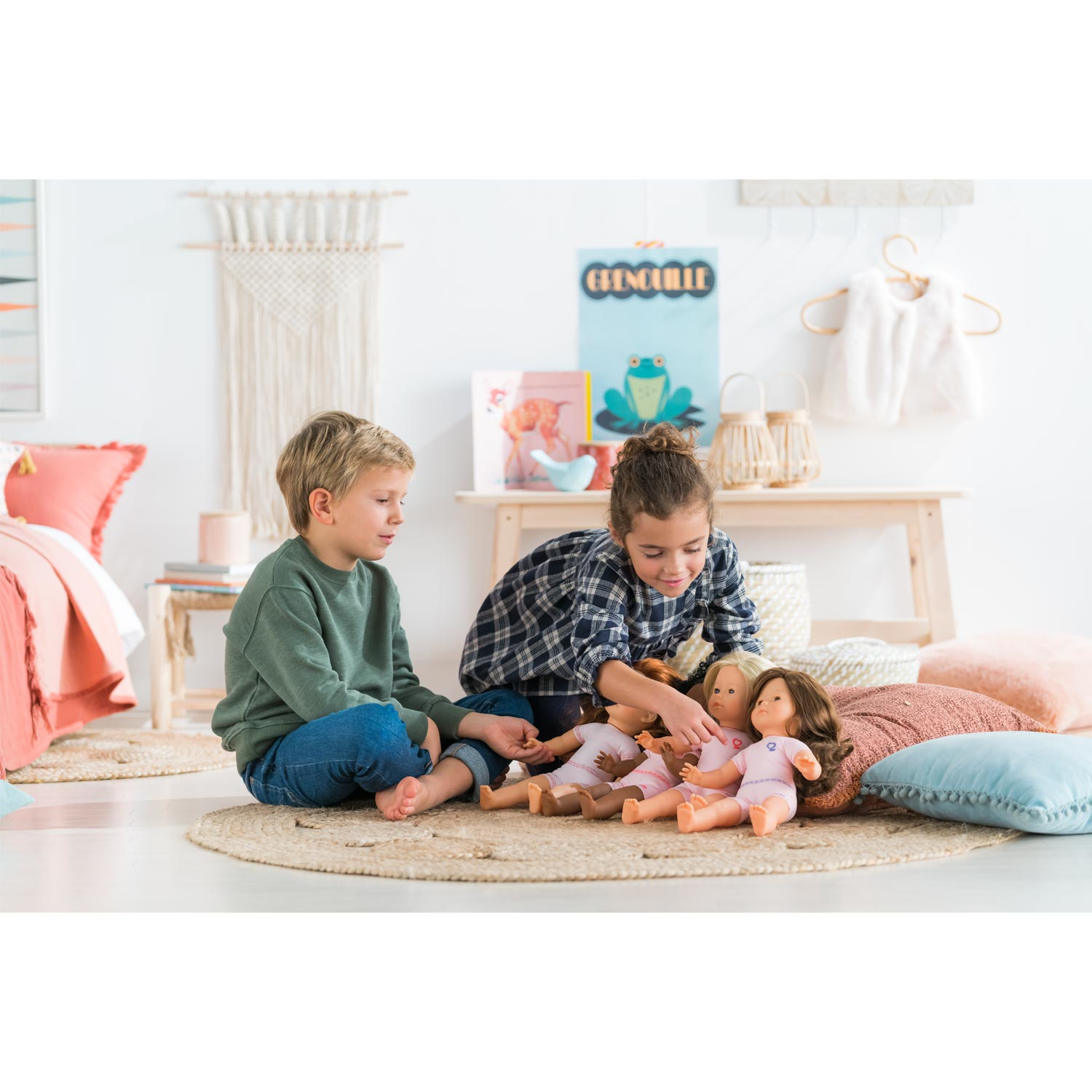 Dwoje dzieci siedzi na dywanie, bawiąc się lalkami. W pokoju znajduje się łóżko, stół i dekoracje na ścianie.