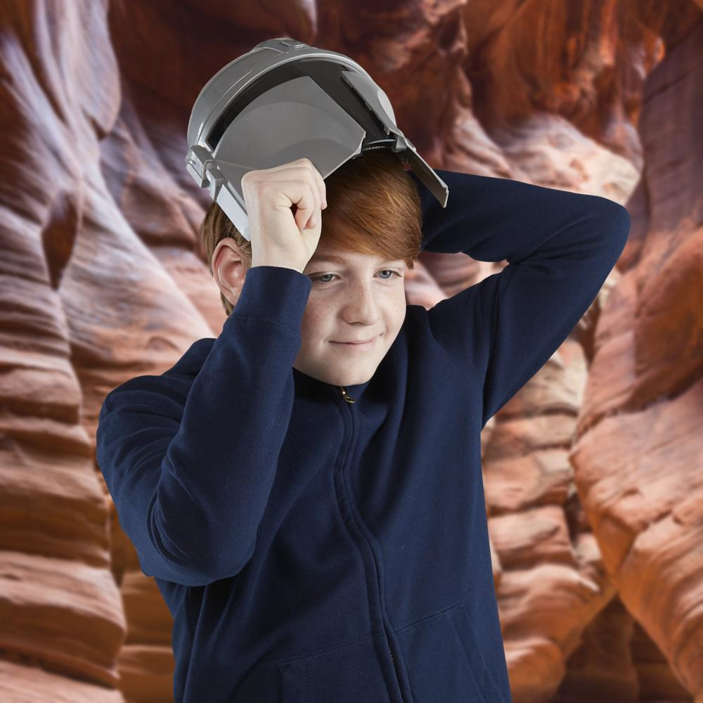 Un niño pelirrojo sostiene un casco gris sobre su cabeza. Lleva una chaqueta azul marino con cremallera. El fondo tiene formaciones rocosas.