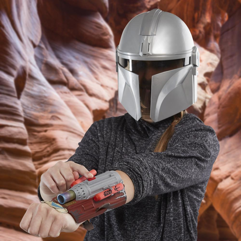 Persona con casco mandaloriano y juguete de brazalete. El brazalete es rojo y gris, y el fondo es un cañón.
