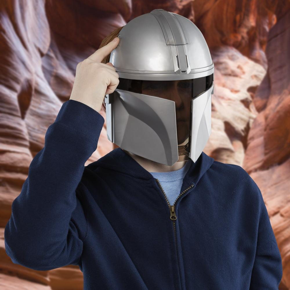 Una persona con un casco plateado de Mandaloriano, tocándolo con un dedo. Lleva una sudadera con capucha azul. Fondo de formación rocosa marrón.