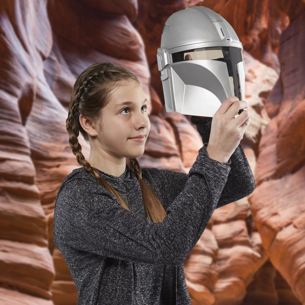 Una niña sostiene un casco mandaloriano. Está frente a un cañón. El casco es plateado, y la niña tiene trenzas, usando una camisa gris.