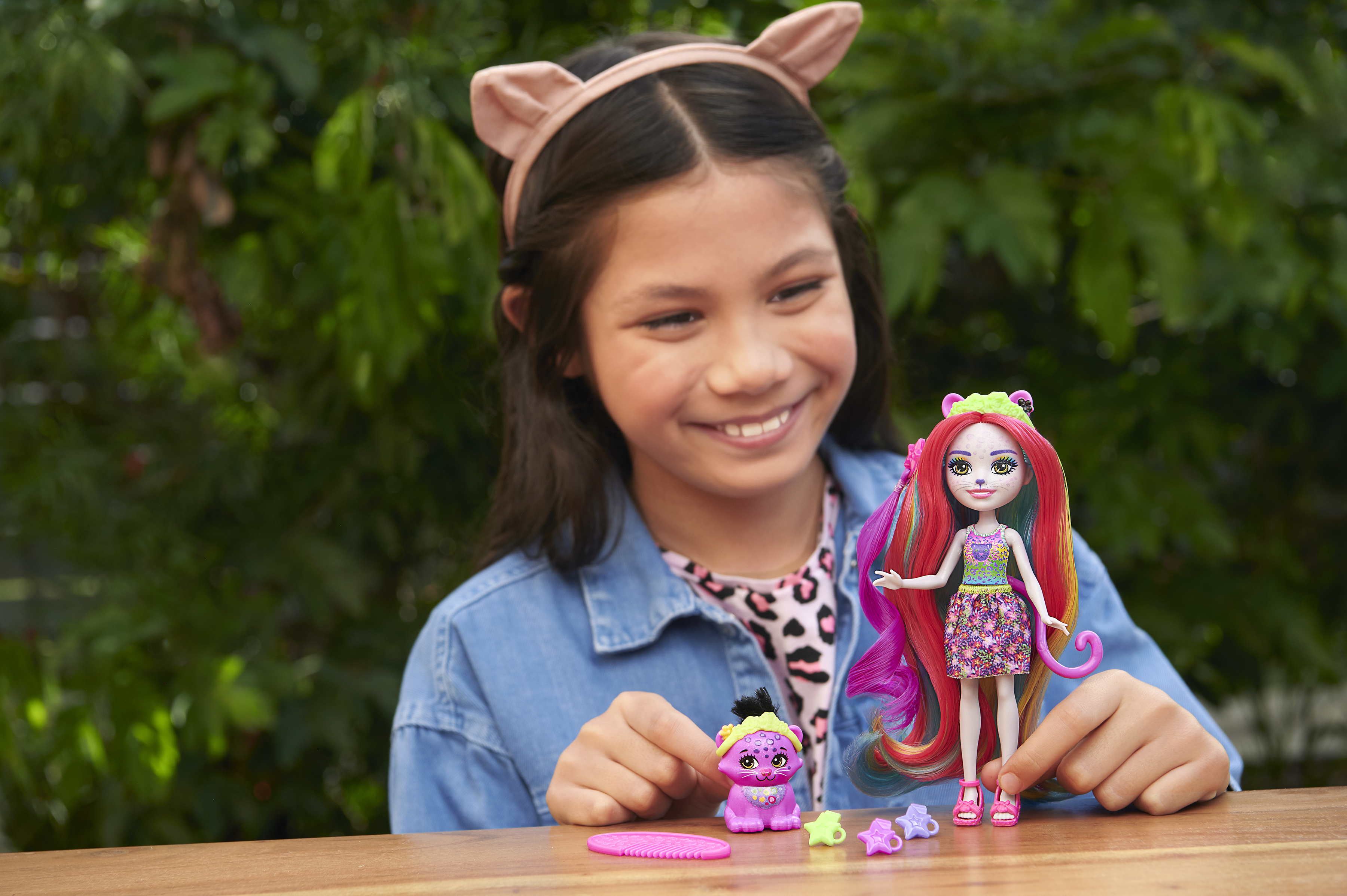 Dziewczyna uśmiecha się, trzymając lalkę. Lalka ma rude włosy i stoi obok zabawkowego zwierzęcia. Zielone tło.