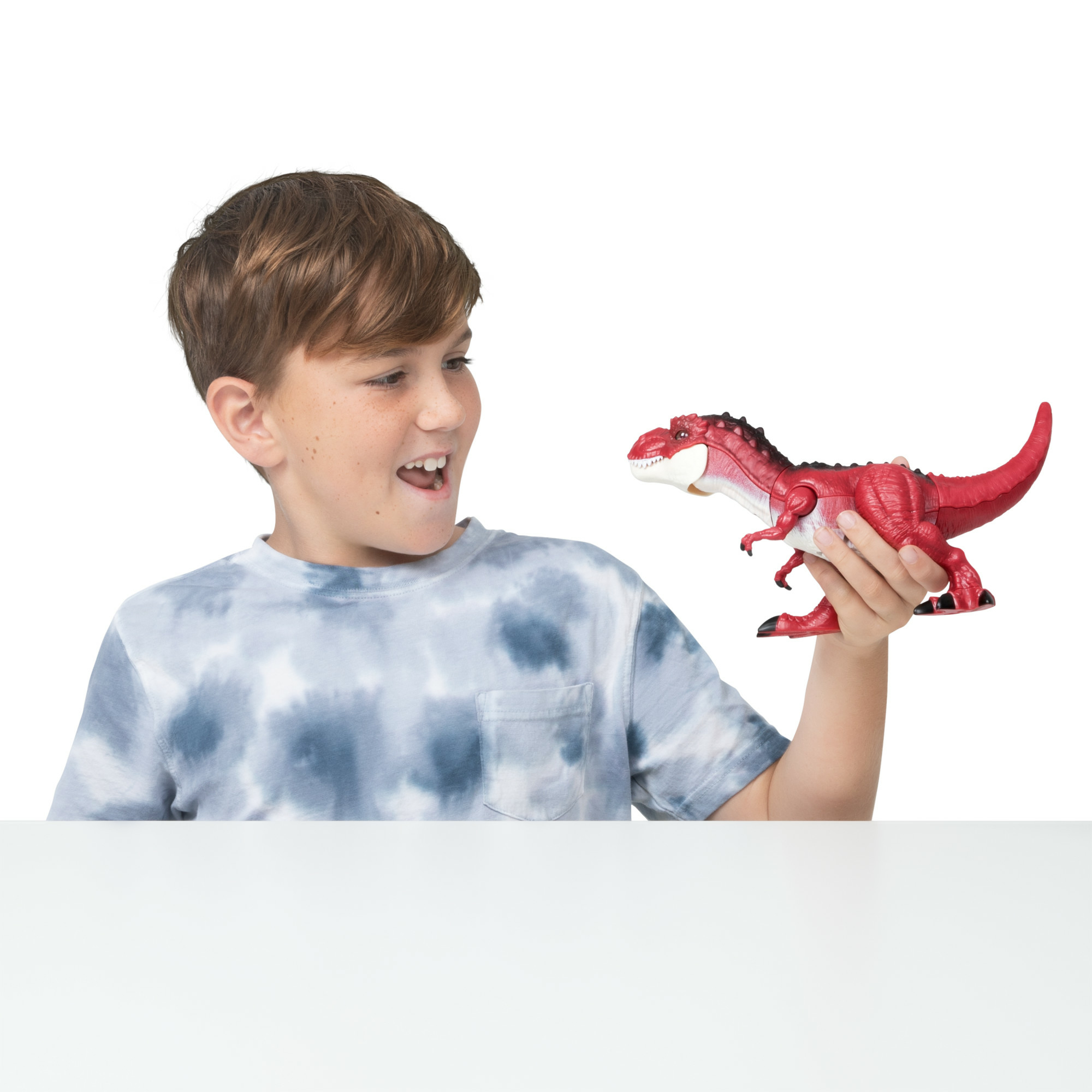 Un niño sonríe a un dinosaurio de juguete rojo. Lleva una camisa teñida. El fondo es blanco.