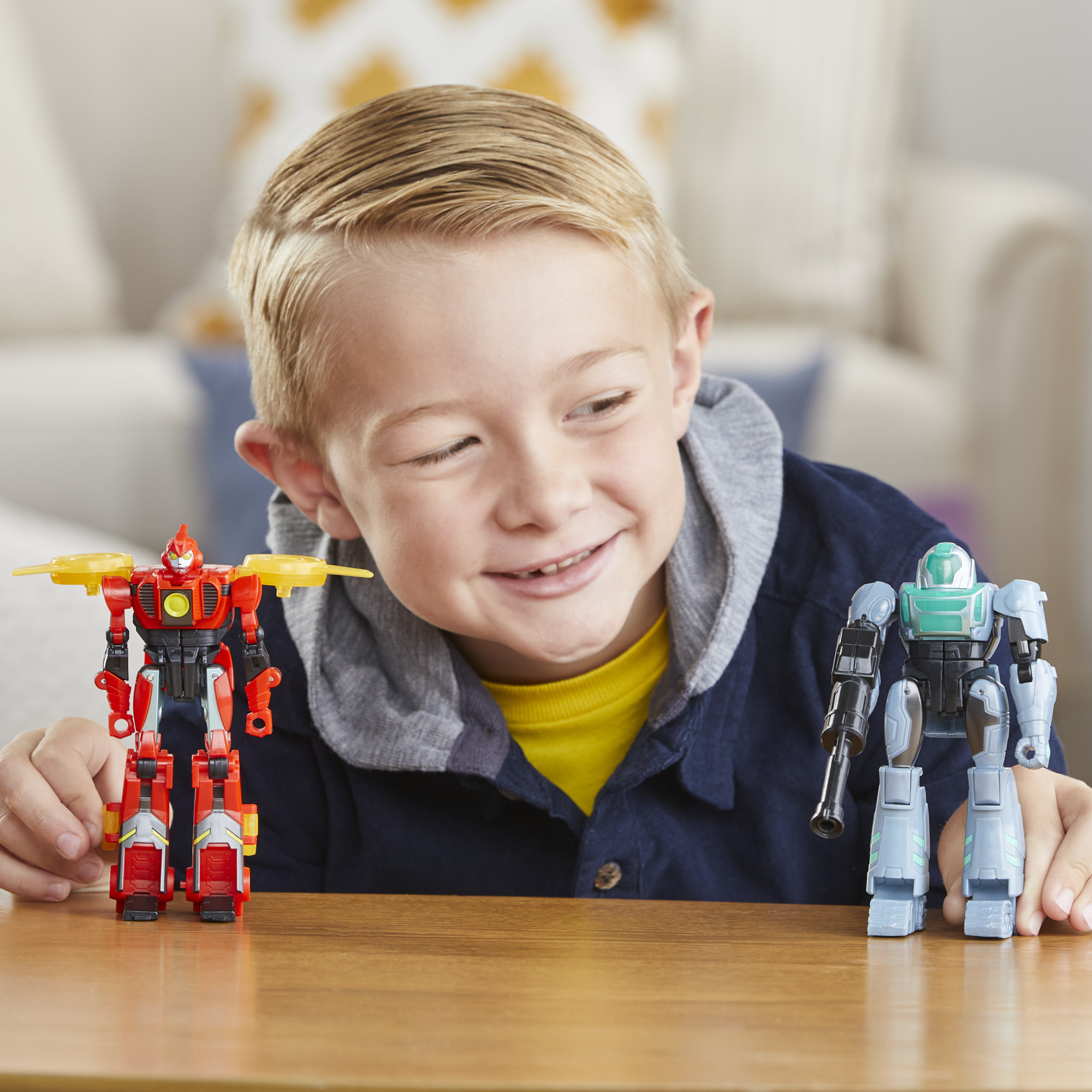 Un ragazzo sorride, guardando due robot giocattolo. Uno rosso, uno blu. Sono su un tavolo di legno. Lo sfondo è un soggiorno.