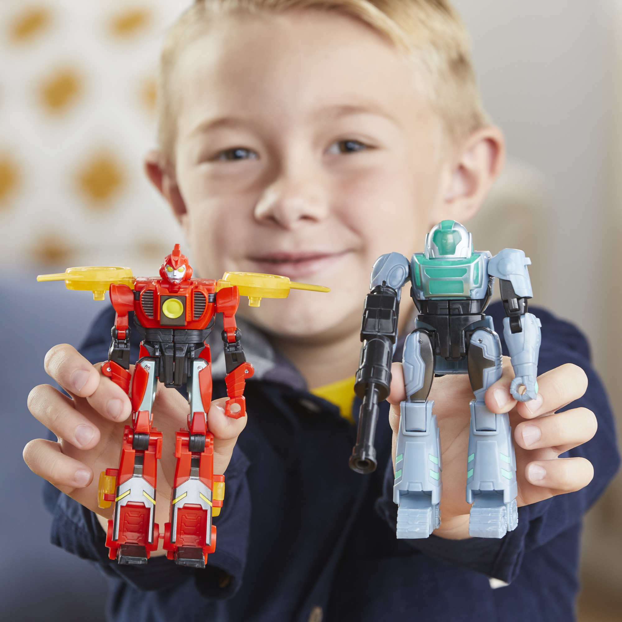 Un ragazzo tiene in mano due giocattoli robot. Uno è rosso, giallo. L'altro è blu, grigio e nero. Il ragazzo sorride alla fotocamera.