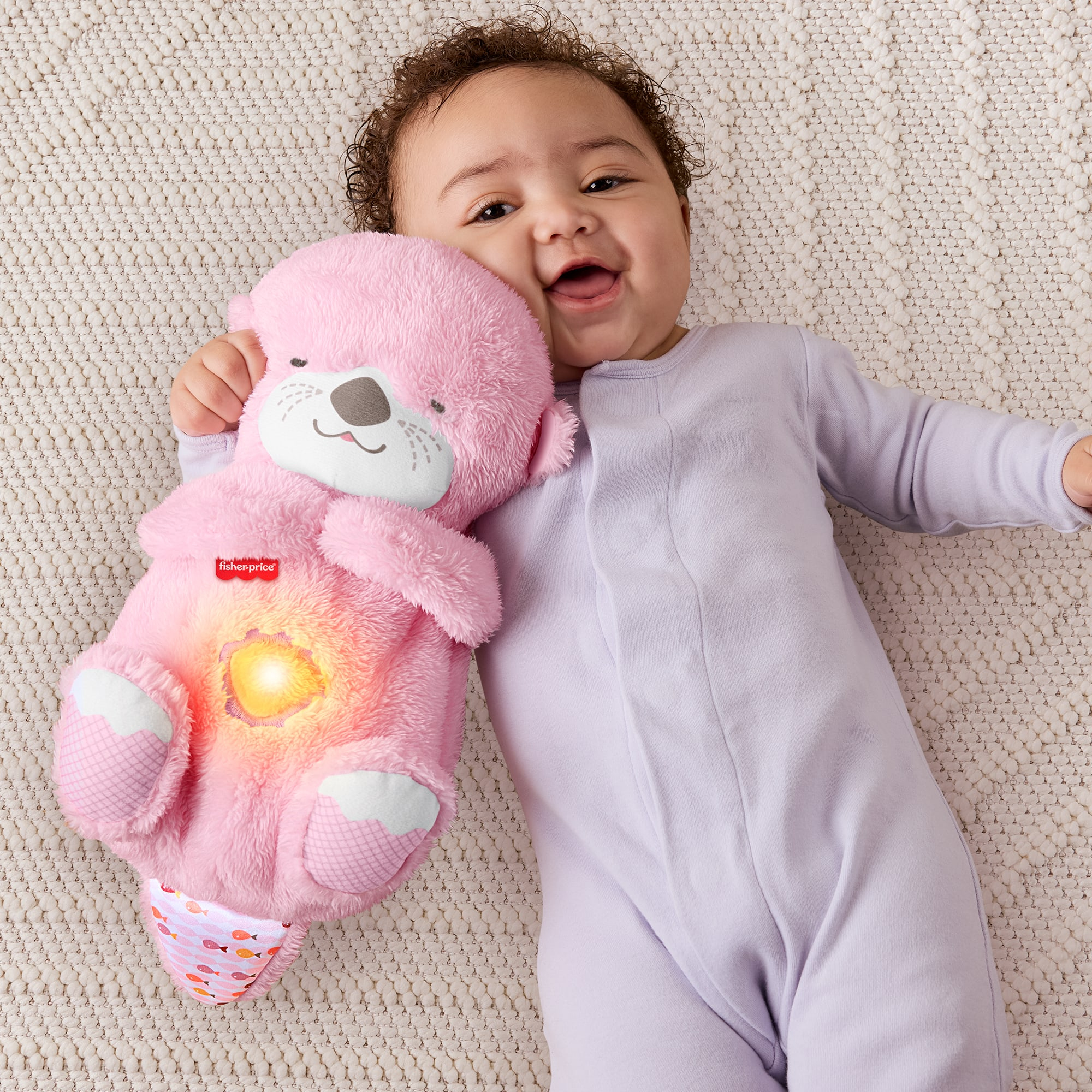Ein Baby lächelt, während es ein rosa Otter-Plüschtier mit leuchtender Mitte hält, das auf einer strukturierten weißen Decke liegt.