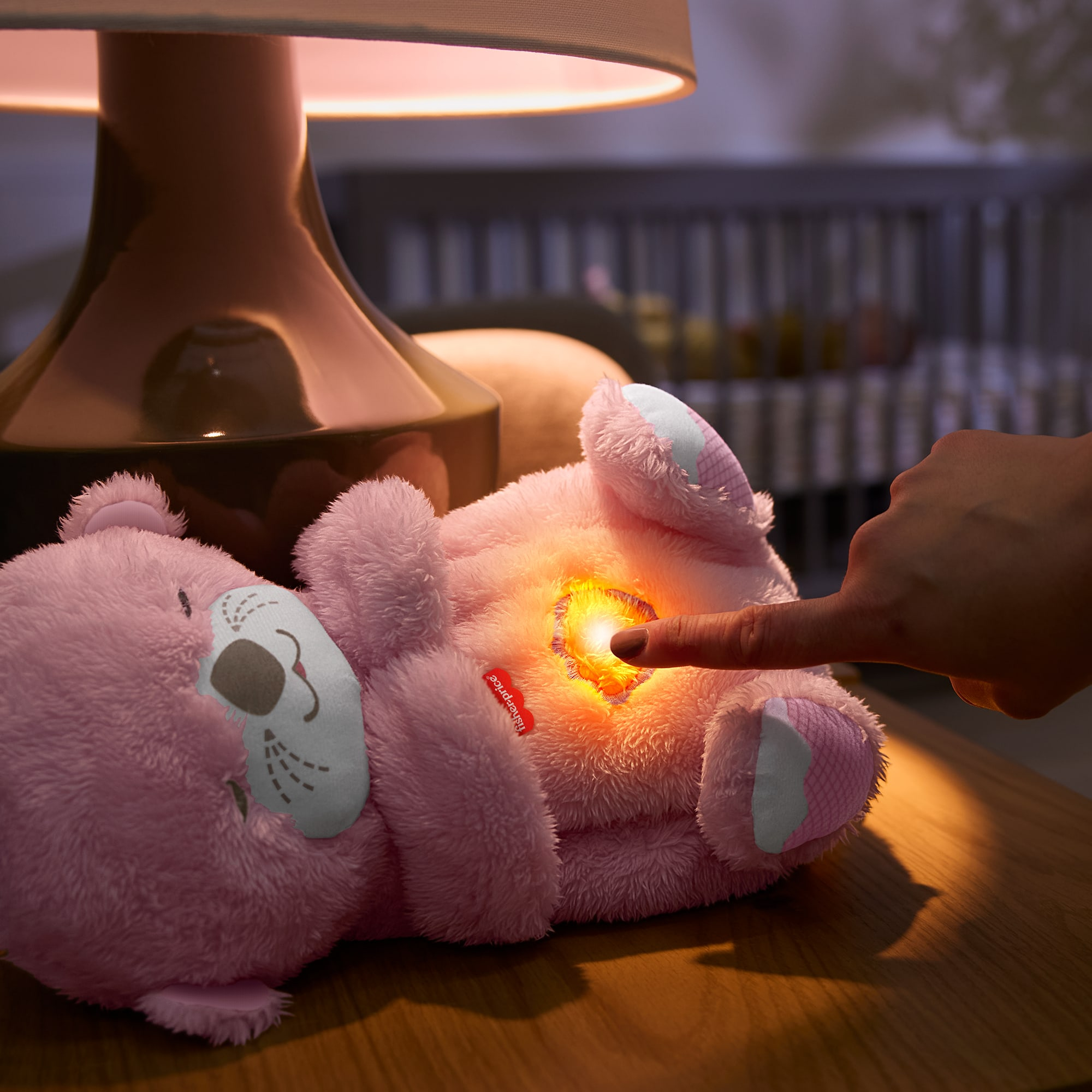 Ein rosa Teddybär-Spielzeug mit leuchtendem Zentrum, ein Finger zeigt darauf. Oben ist eine Lampe. Im Hintergrund ist ein Kinderbett.
