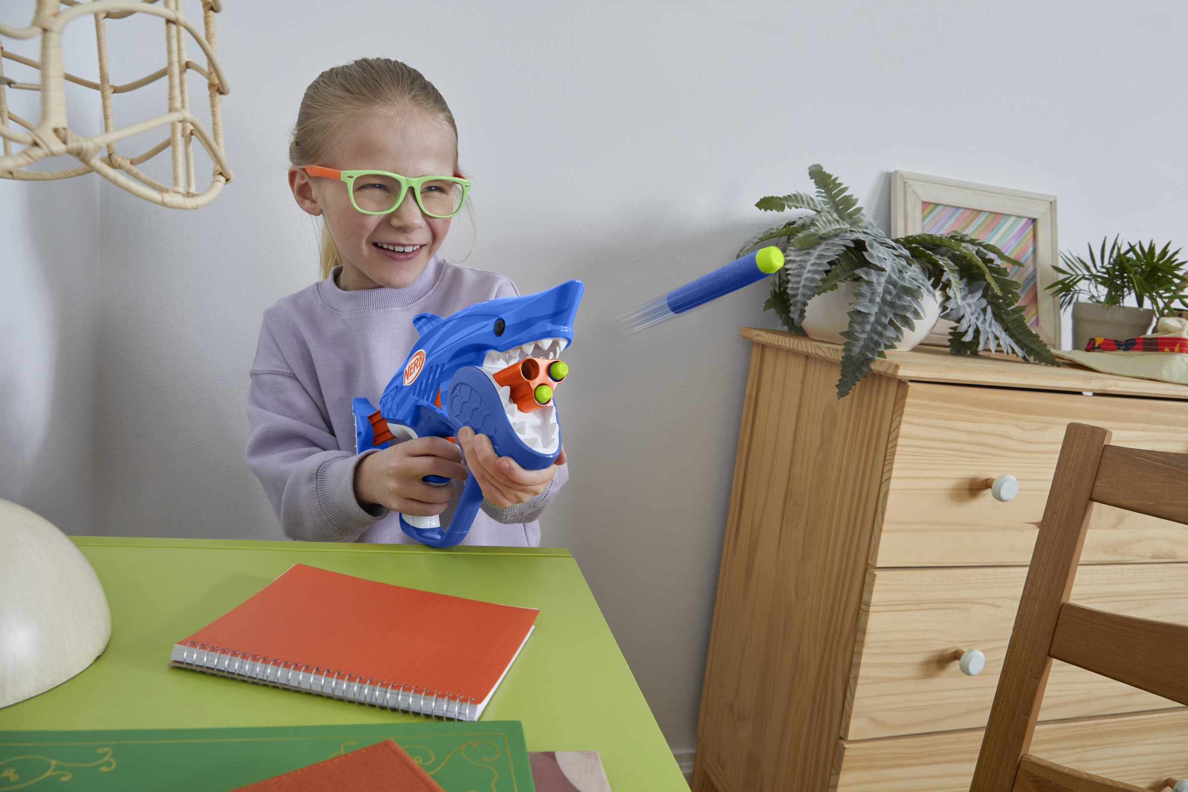 Dziewczyna w okularach strzela z zabawkowego pistoletu w kształcie rekina. Strzałka leci. W tle biurko, rośliny i komoda.