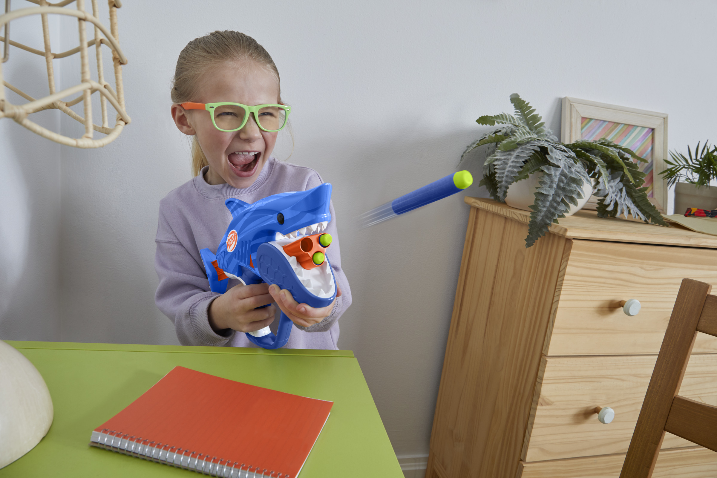 Dziewczynka w okularach strzela z niebieskiej zabawki w kształcie rekina. Pocisk jest w ruchu. Notatnik leży na stole.
