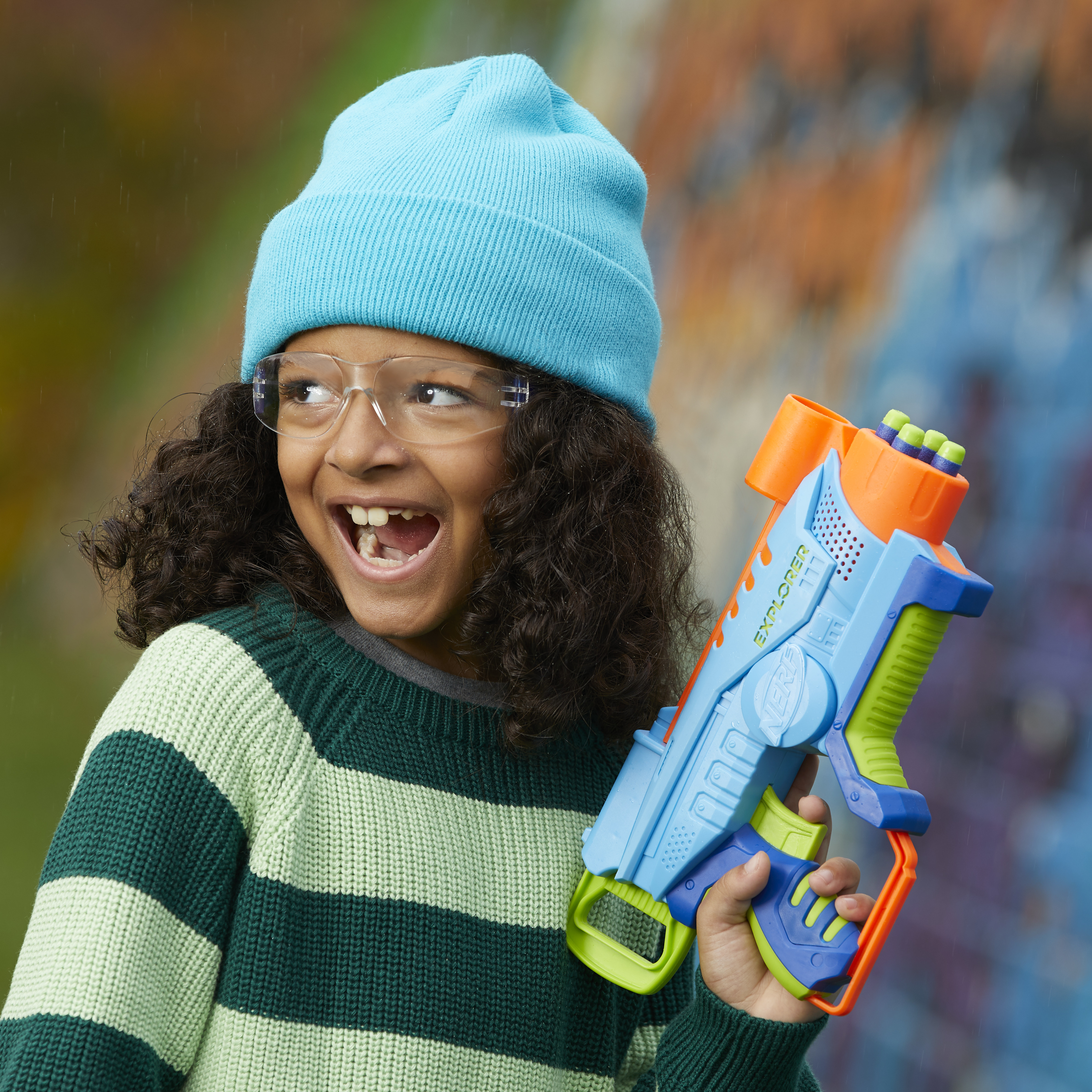 Un niño con gafas y gorro azul sostiene una pistola de juguete, sonriendo. El fondo es un mural colorido.