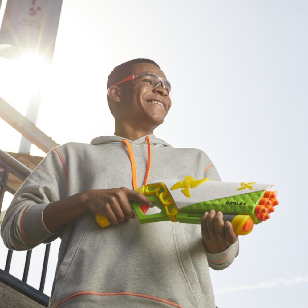 Uśmiechnięty chłopiec w okularach trzyma zabawkę Roblox. Ma na sobie szarą bluzę z kapturem. Tło obejmuje budynek i jasne niebo.