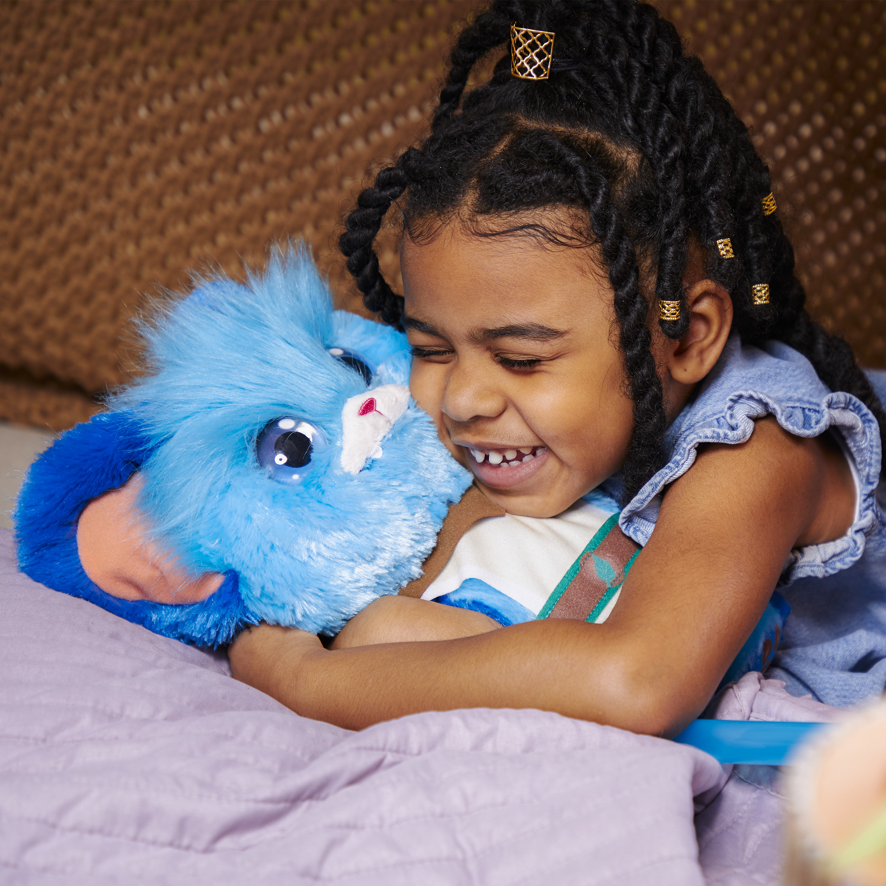 Uśmiechnięta czarnoskóra dziewczynka przytula niebieską pluszową zabawkę. Ma na sobie dżinsowy top. Złote spinki do włosów są w warkoczach.