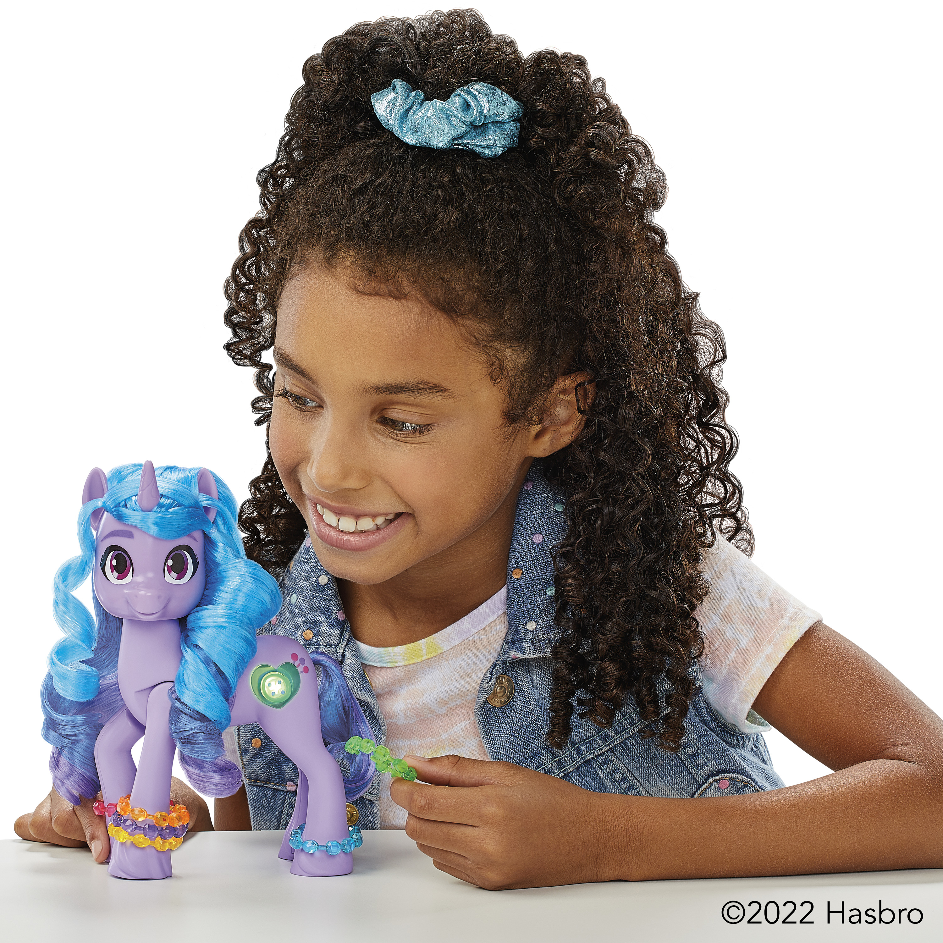 Una niña sonríe a un poni de juguete. El poni es morado con pelo azul. La niña lleva un chaleco vaquero y tiene el pelo rizado.