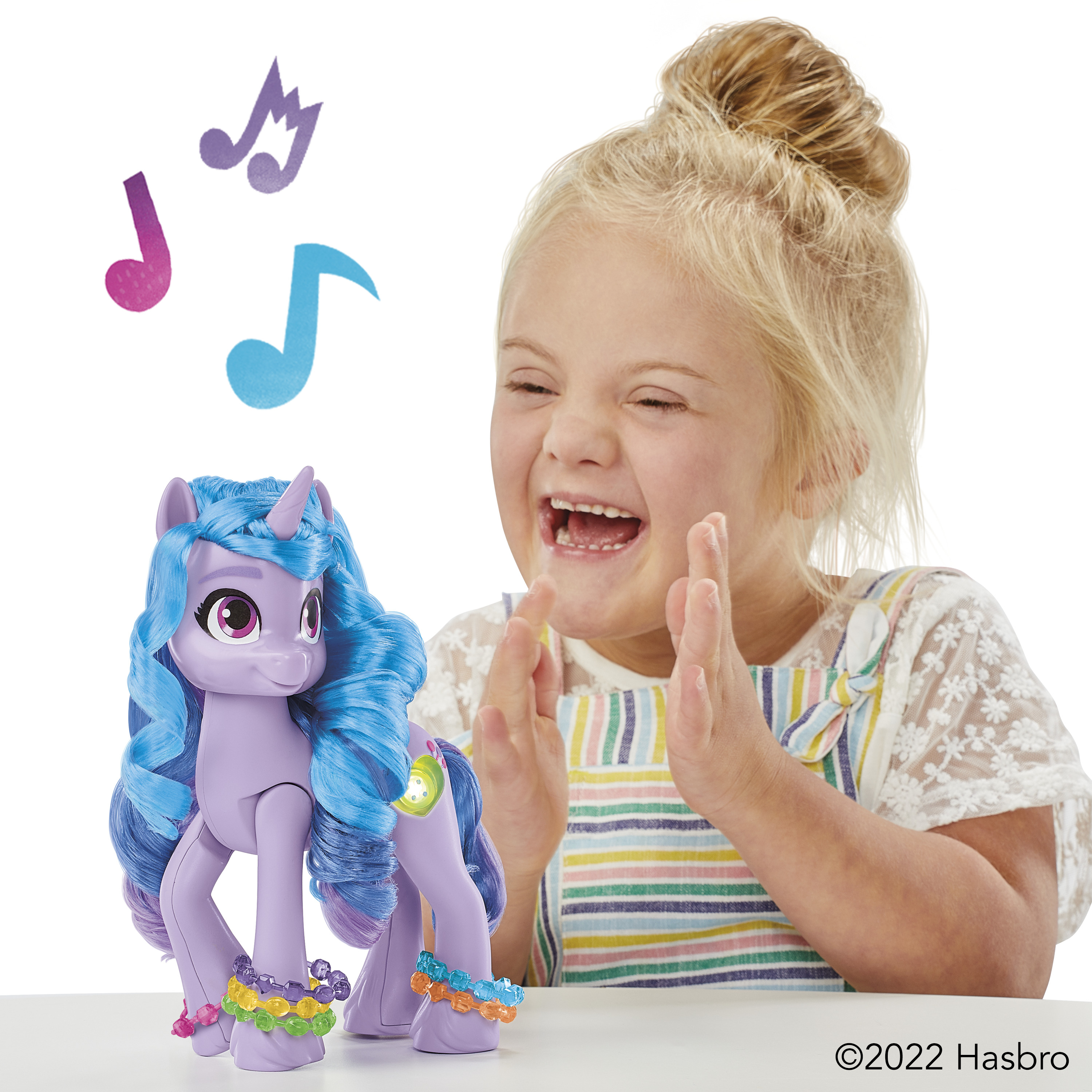 Una niña se ríe de un poni de juguete. El poni es morado con pelo azul, con pulseras. Notas musicales flotan arriba.