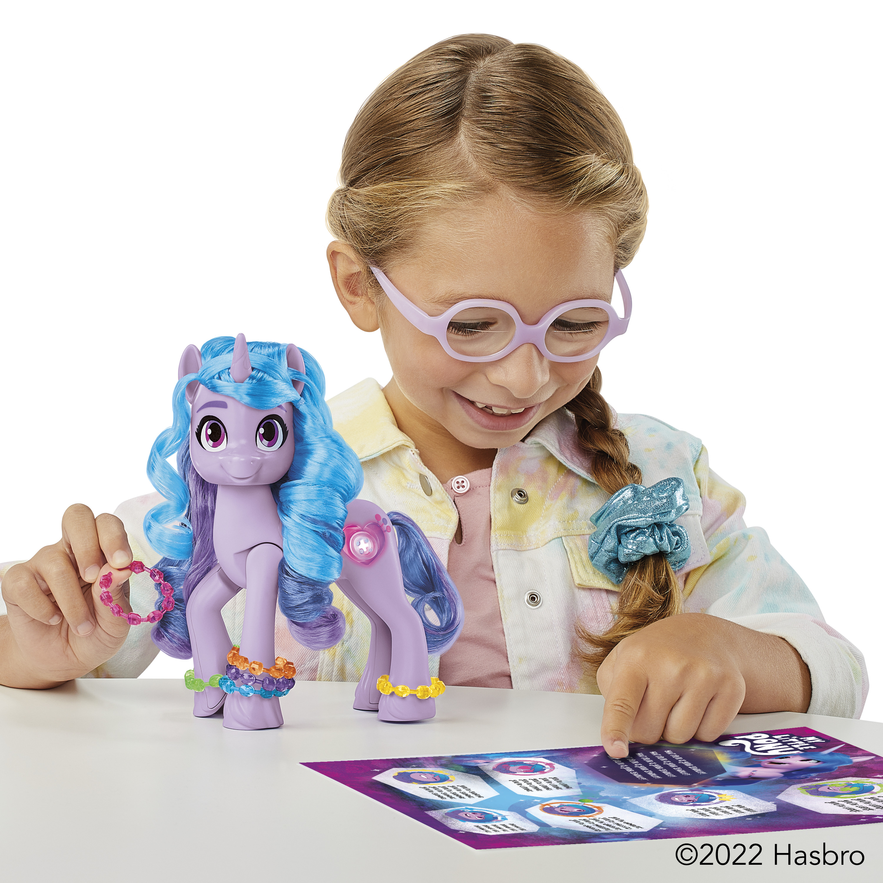 Una niña con gafas juega con un juguete de pony morado. El juguete tiene pelo azul y pulseras coloridas. Hay un juego en la mesa.