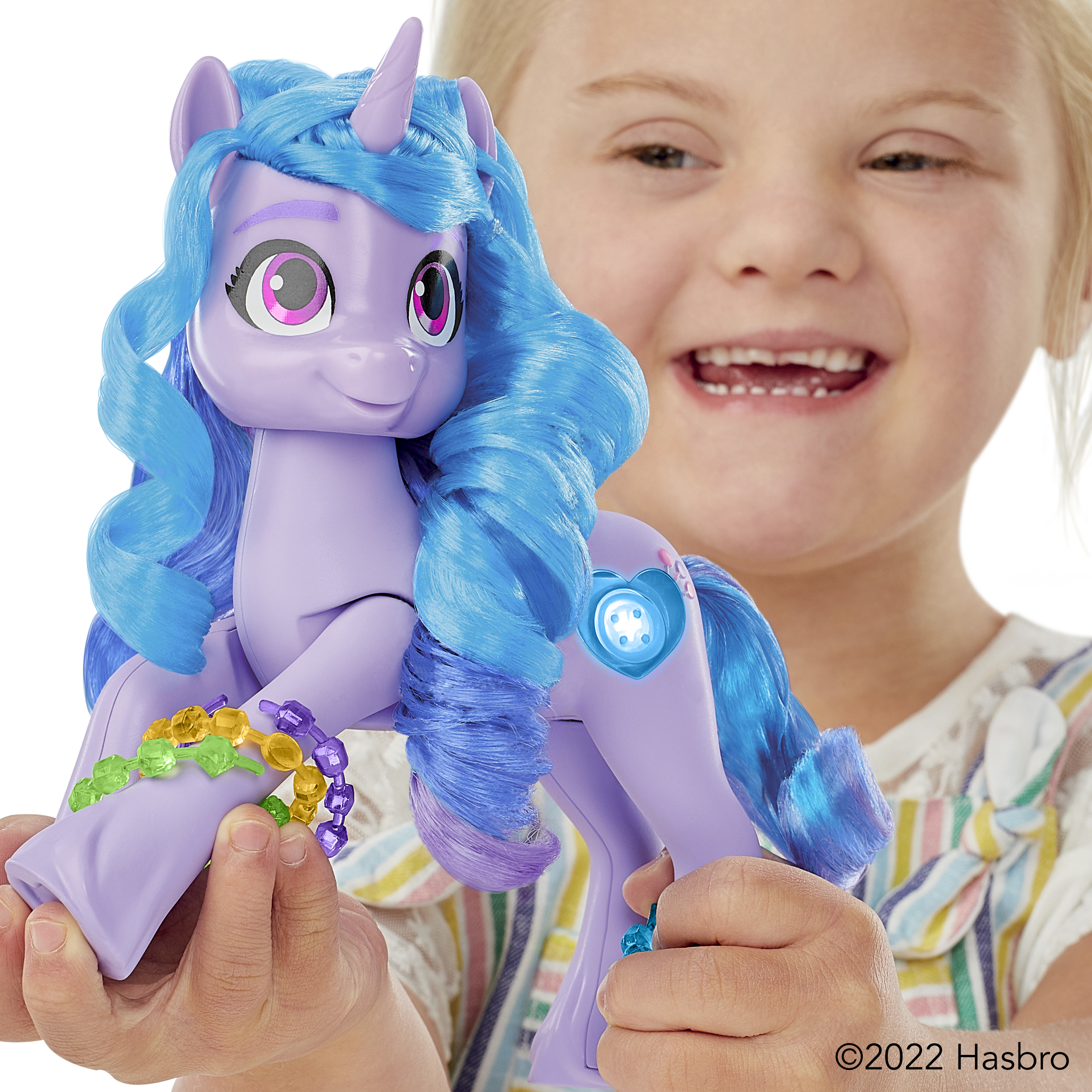 Una niña sostiene un poni de juguete morado con pelo azul. El poni tiene un cuerno, un botón en forma de corazón y una pulsera colorida.