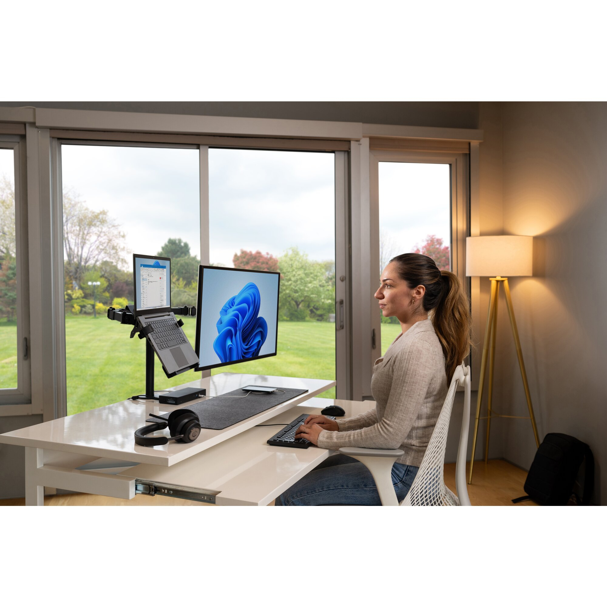 Vrouw aan bureau met dubbele monitoren, laptop en koptelefoon. Windows bureaublad zichtbaar. Groot raam met groen.