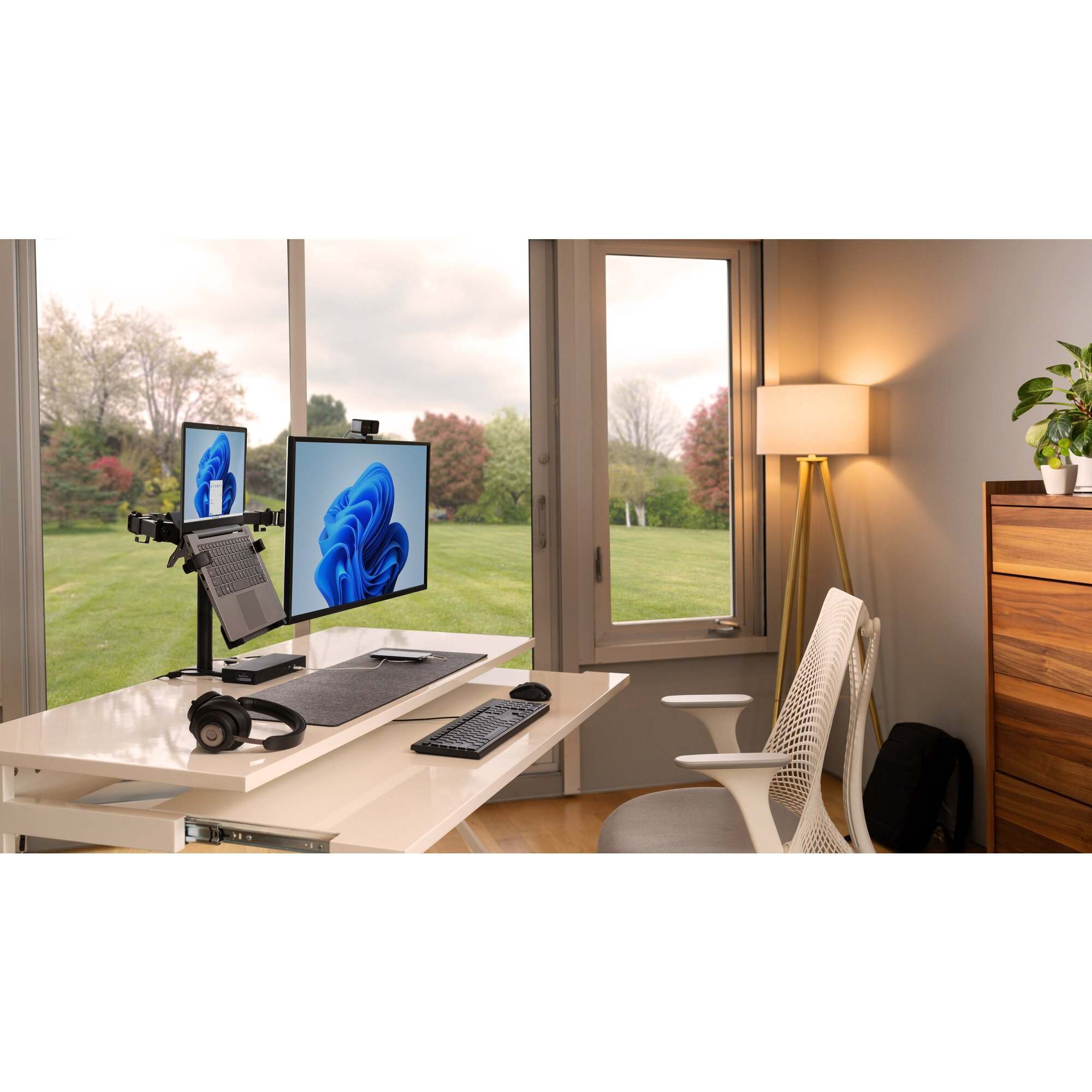 Un moderno allestimento per ufficio con doppio monitor, laptop e cuffie su una scrivania bianca, con vista su un giardino verde.